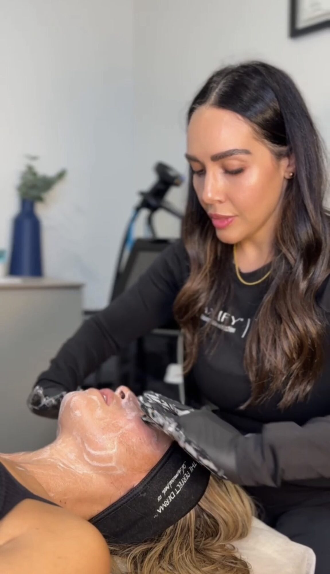 A woman with dark hair and a black shirt performs a facial treatment on a person lying down, in a clinic setting.