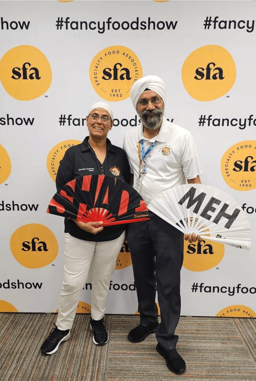 Two men standing in front of a backdrop with logos, holding signs, smiling, one wearing a turban and glasses, the other in a white shirt.