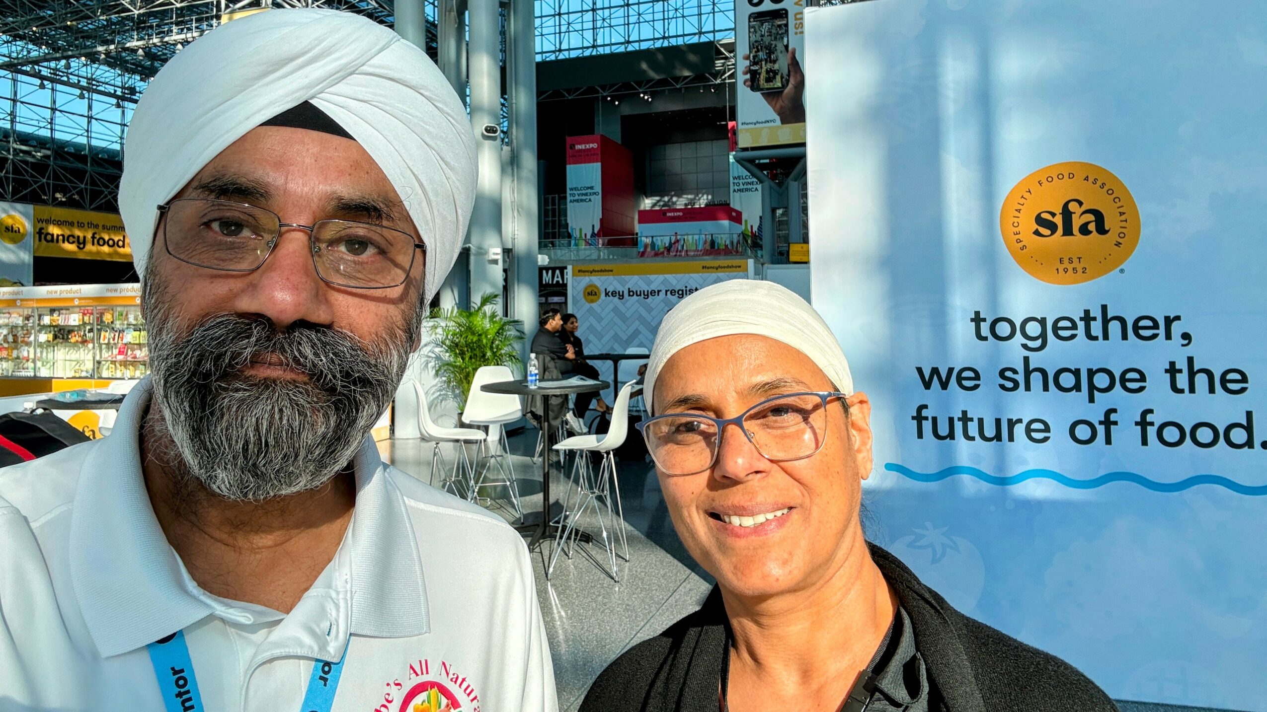 Two people wearing white head coverings and glasses stand outdoors near a food event, smiling, with a blue banner in the background.