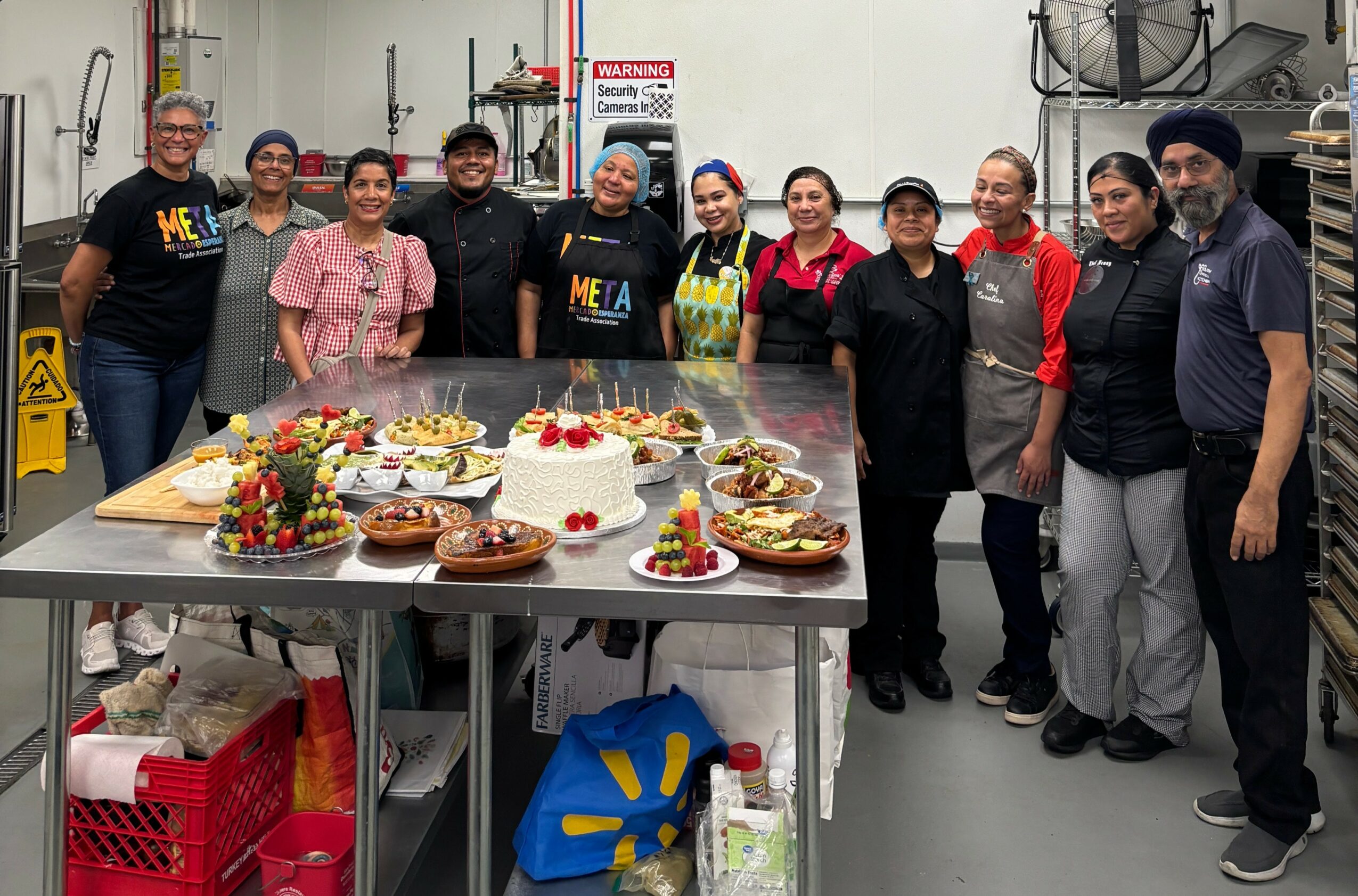 Group of ten people standing behind a table with decorated cakes and desserts in a commercial kitchen.