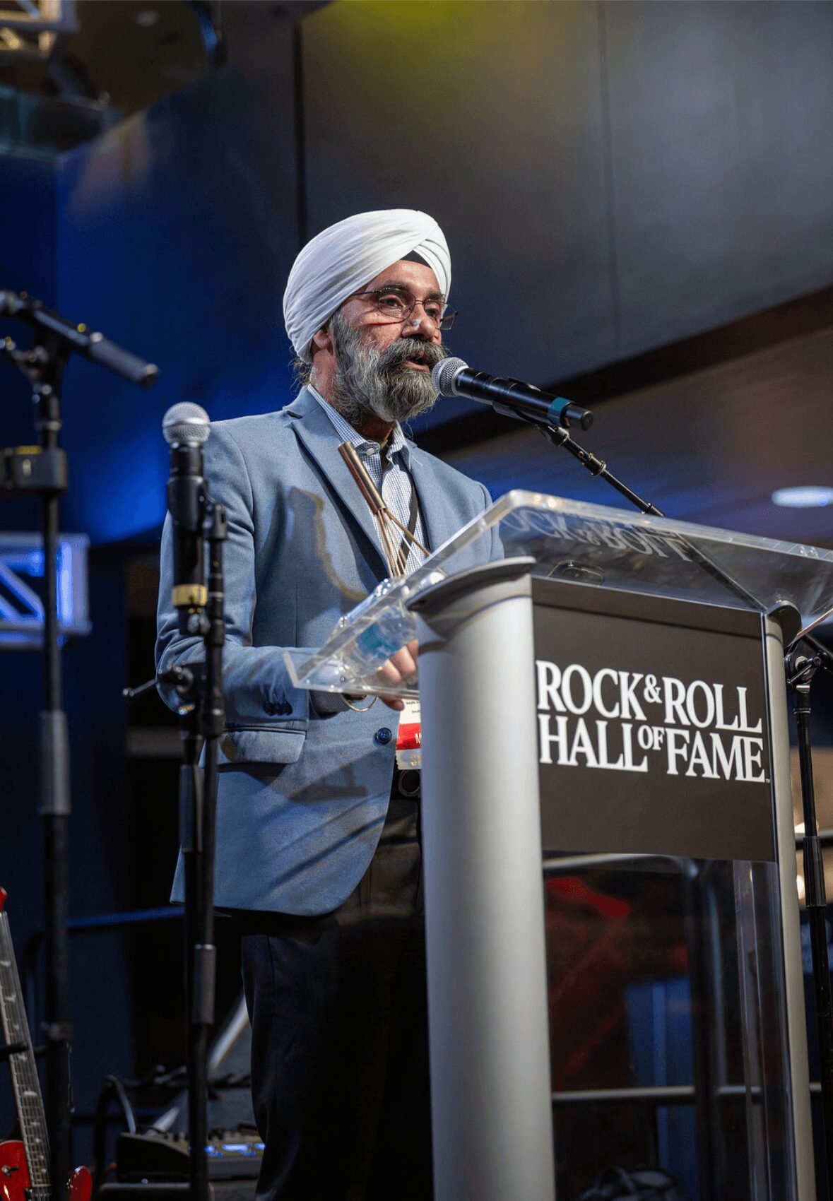 Man wearing a turban and glasses speaking at a podium during an event, with a sign reading 'Rock & Roll Hall of Fame'.