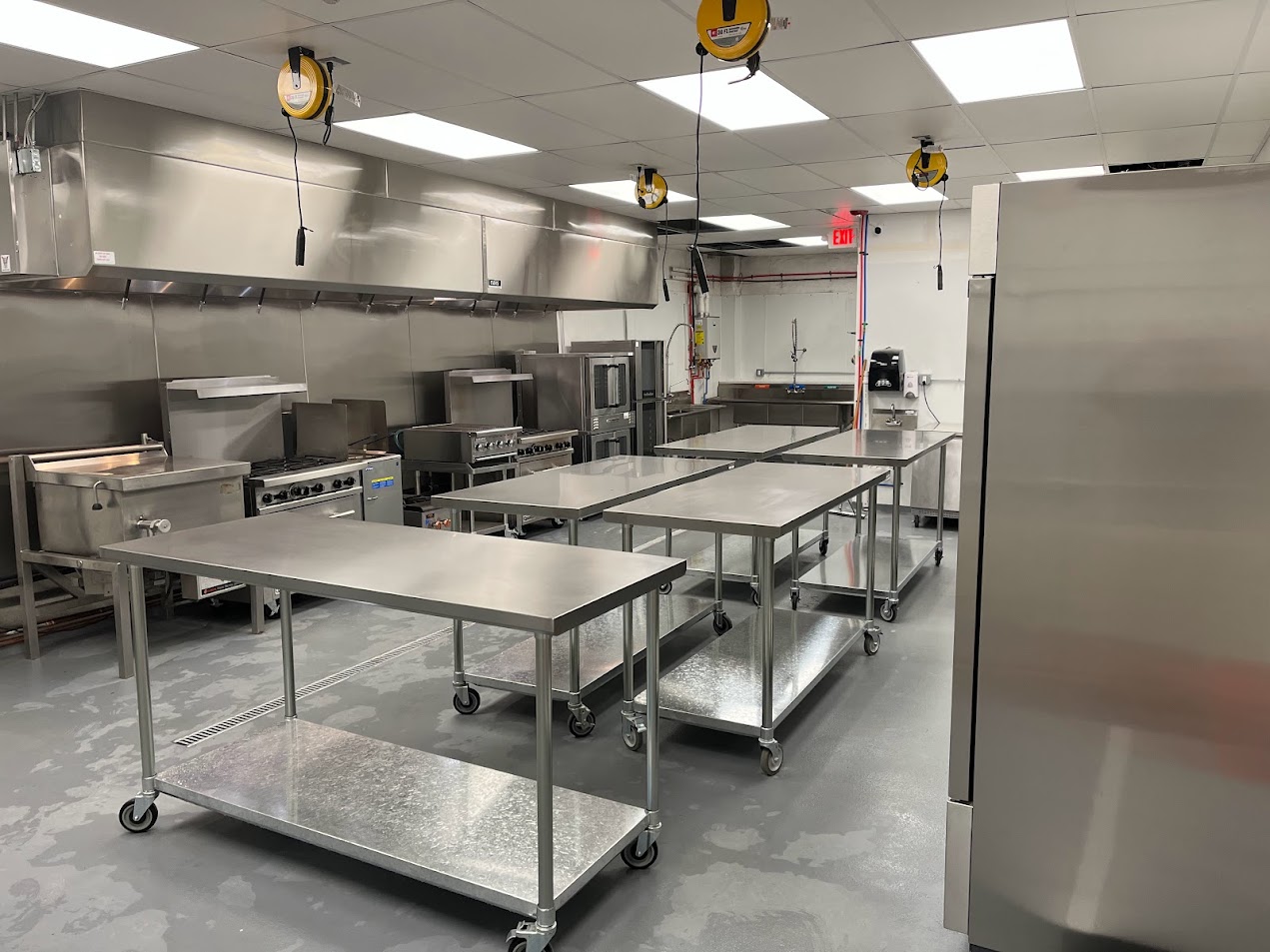 Commercial kitchen with stainless steel tables, appliances, and ceiling lights, empty and clean.