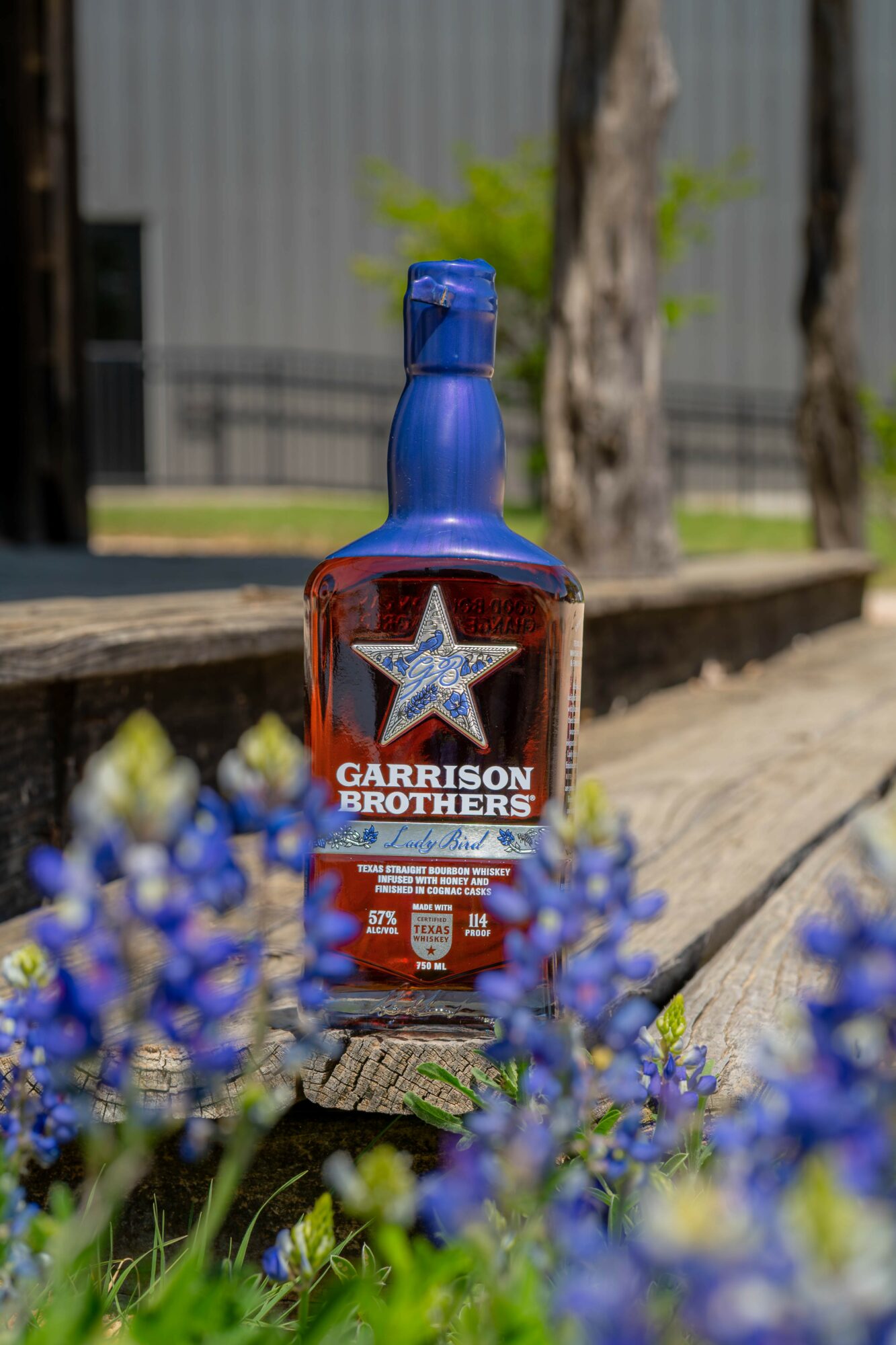 Bottle of Garrison Brothers whiskey on a wooden surface outdoors, with blue flowers in foreground and trees in background.