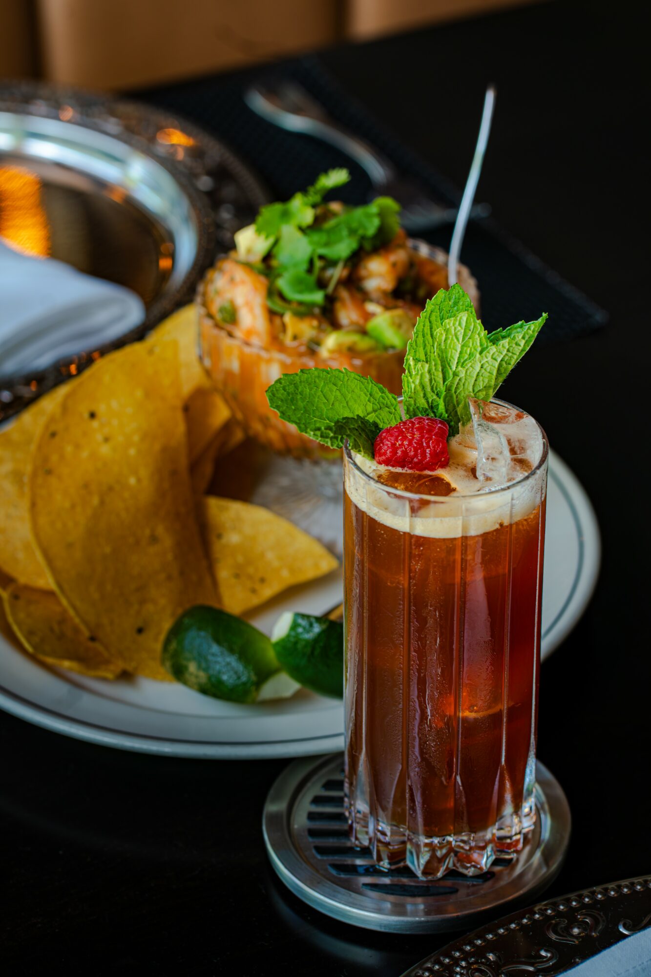 A tall glass of pink cocktail garnished with mint, raspberry, and lime, with a straw, on a table.