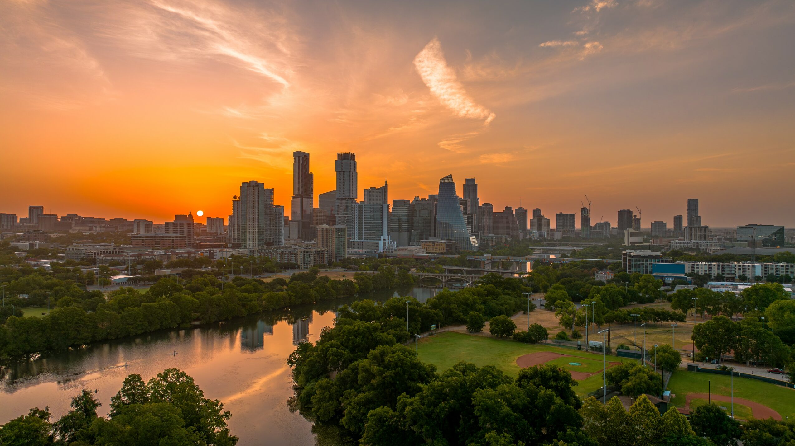 City skyline at sunset with tall buildings, a river, and green park area in foreground.