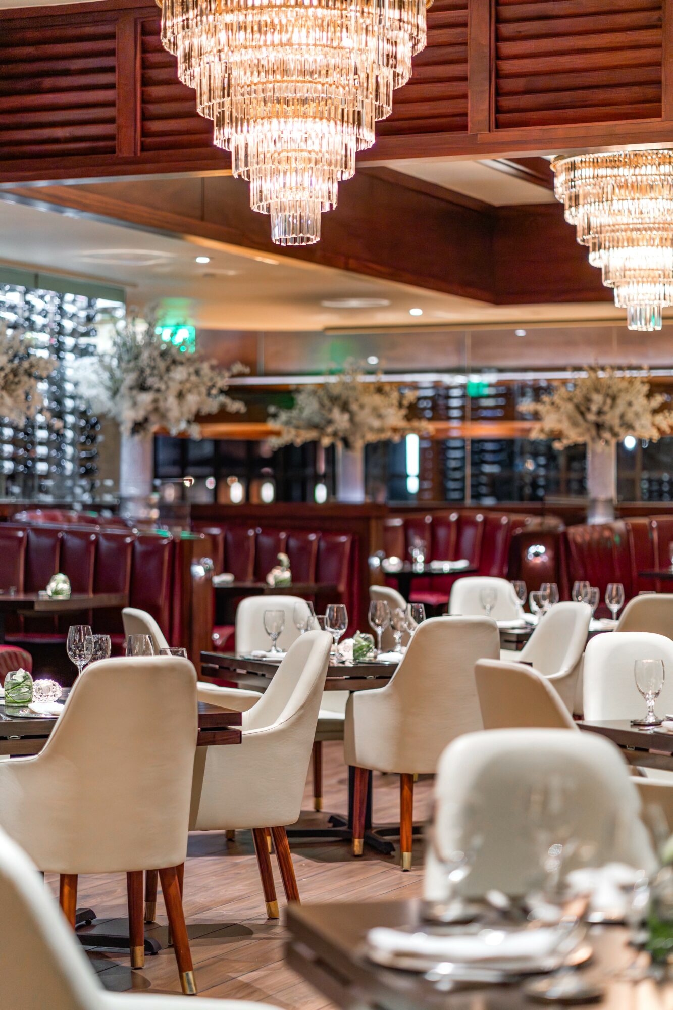 Elegant restaurant interior with chandeliers, white and beige chairs, and tables set for dining, decorated with flowers.