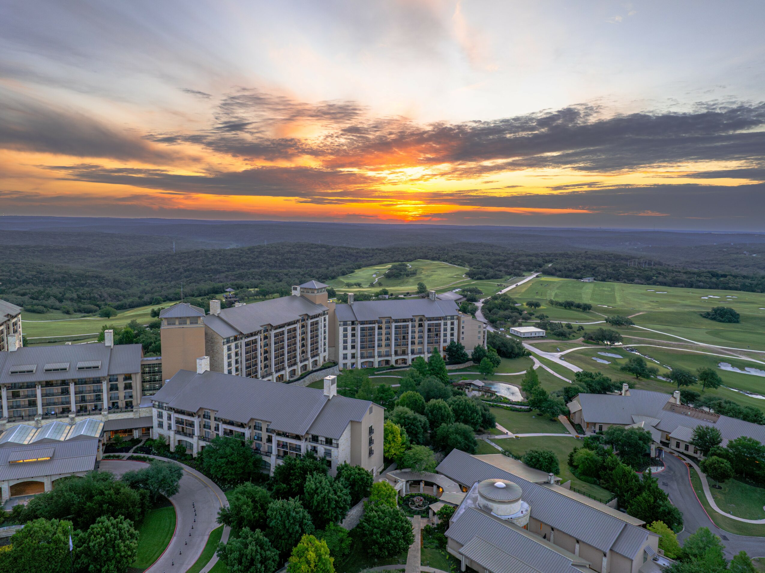 Aerial view of a large building complex with surrounding greenery and open fields at sunset.