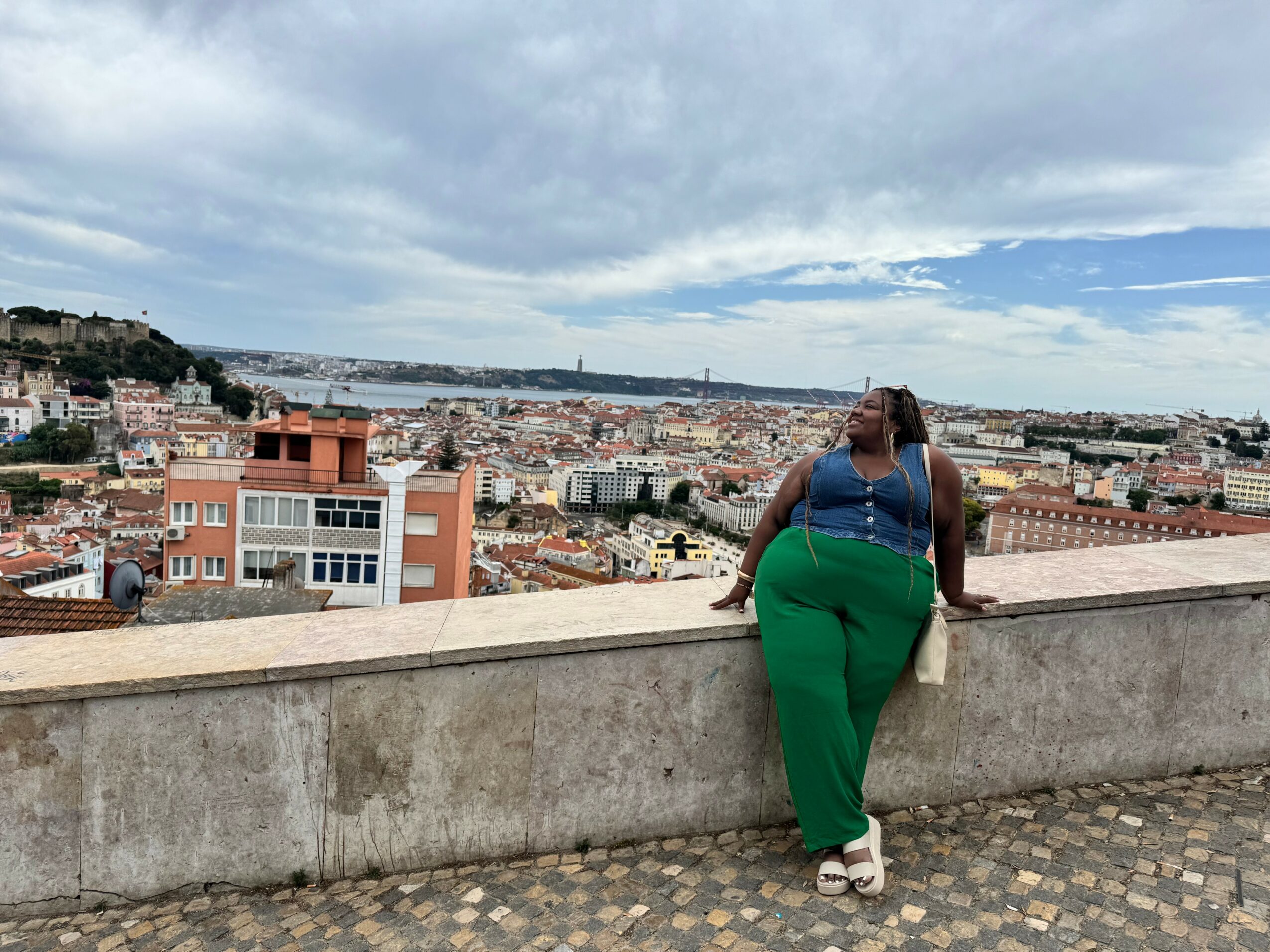 Person with long hair wearing a blue top and green pants leaning on a stone wall overlooking a cityscape and water, cloudy sky.