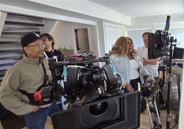Group of people on a film set with cameras and equipment indoors, some looking at monitors, others talking.