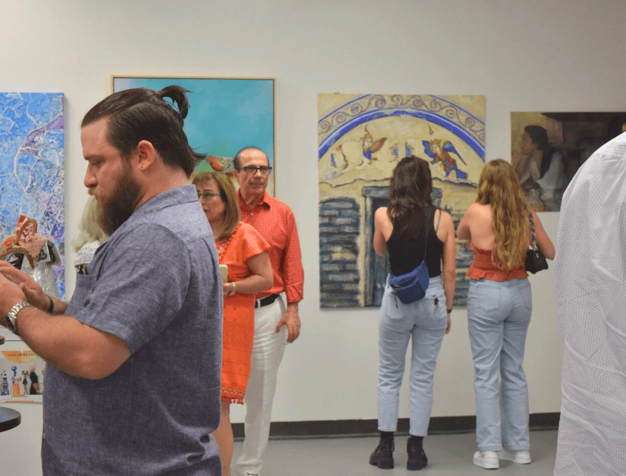 People viewing artwork in an art gallery, some looking at paintings on the wall, others taking photos or talking.