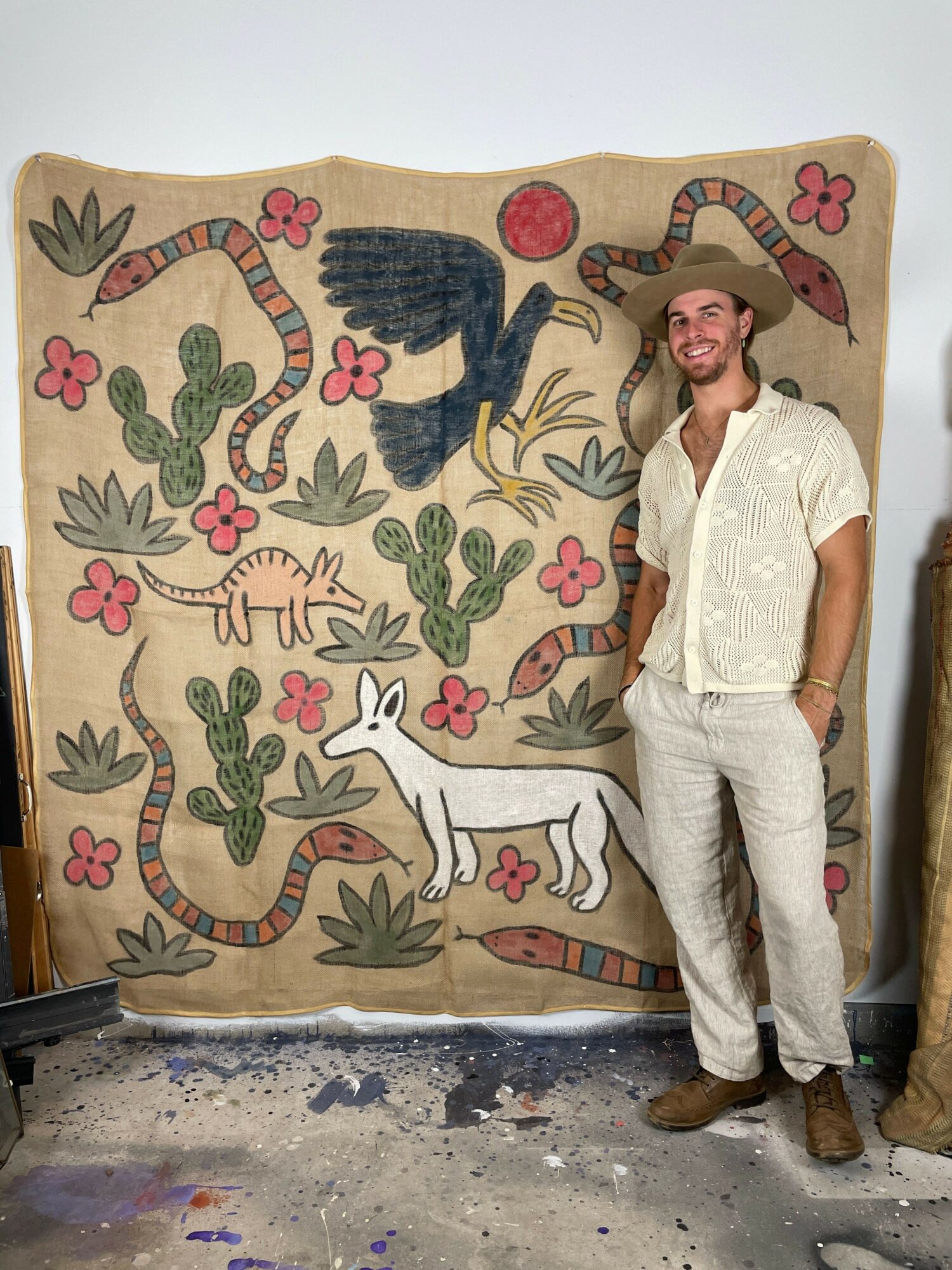 Person standing next to a large, colorful mural with animals and plants, smiling, wearing light-colored clothing.