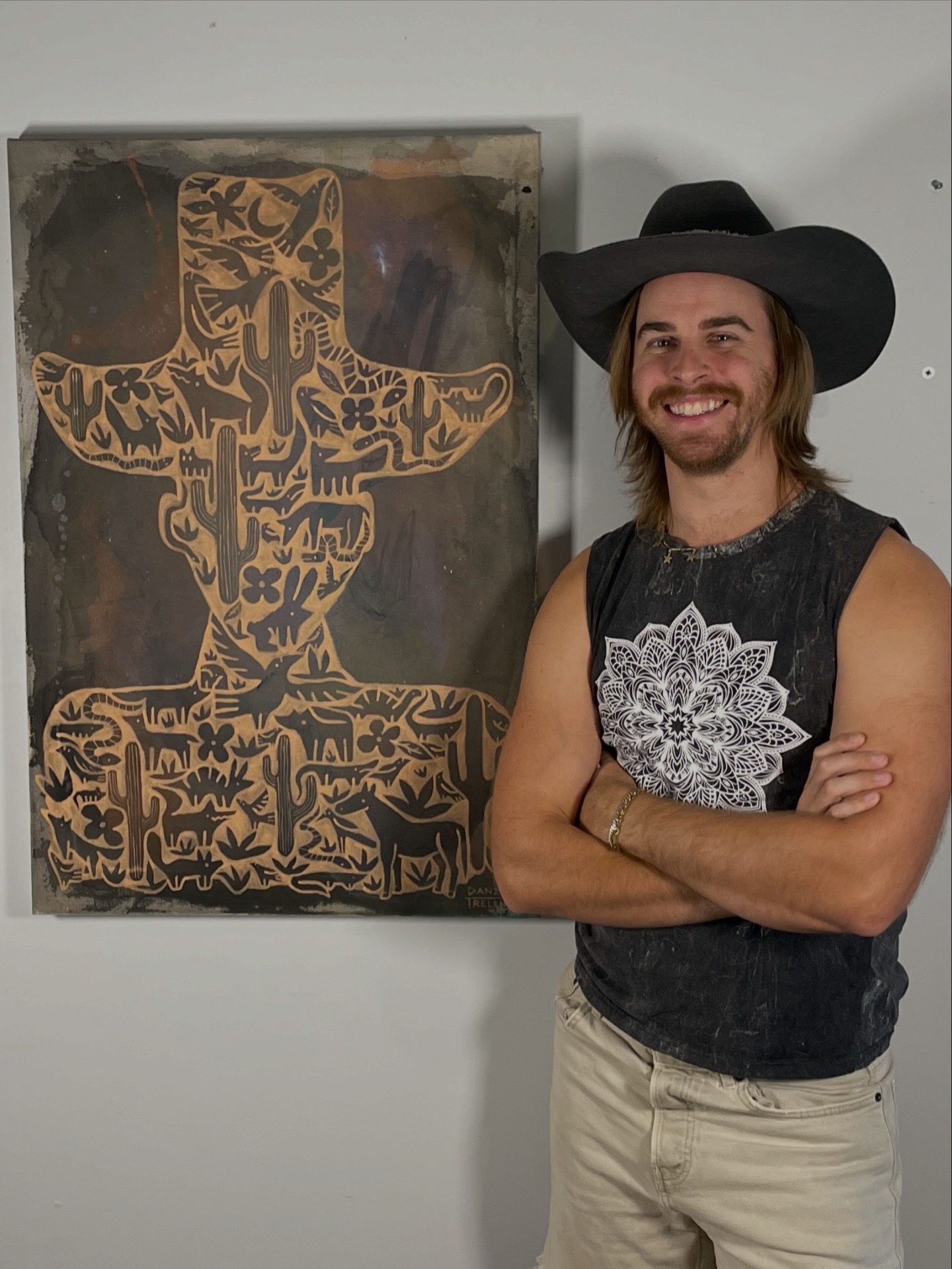 Person with long hair, beard, black hat, sleeveless shirt, and light pants standing with arms crossed, smiling, next to artwork.