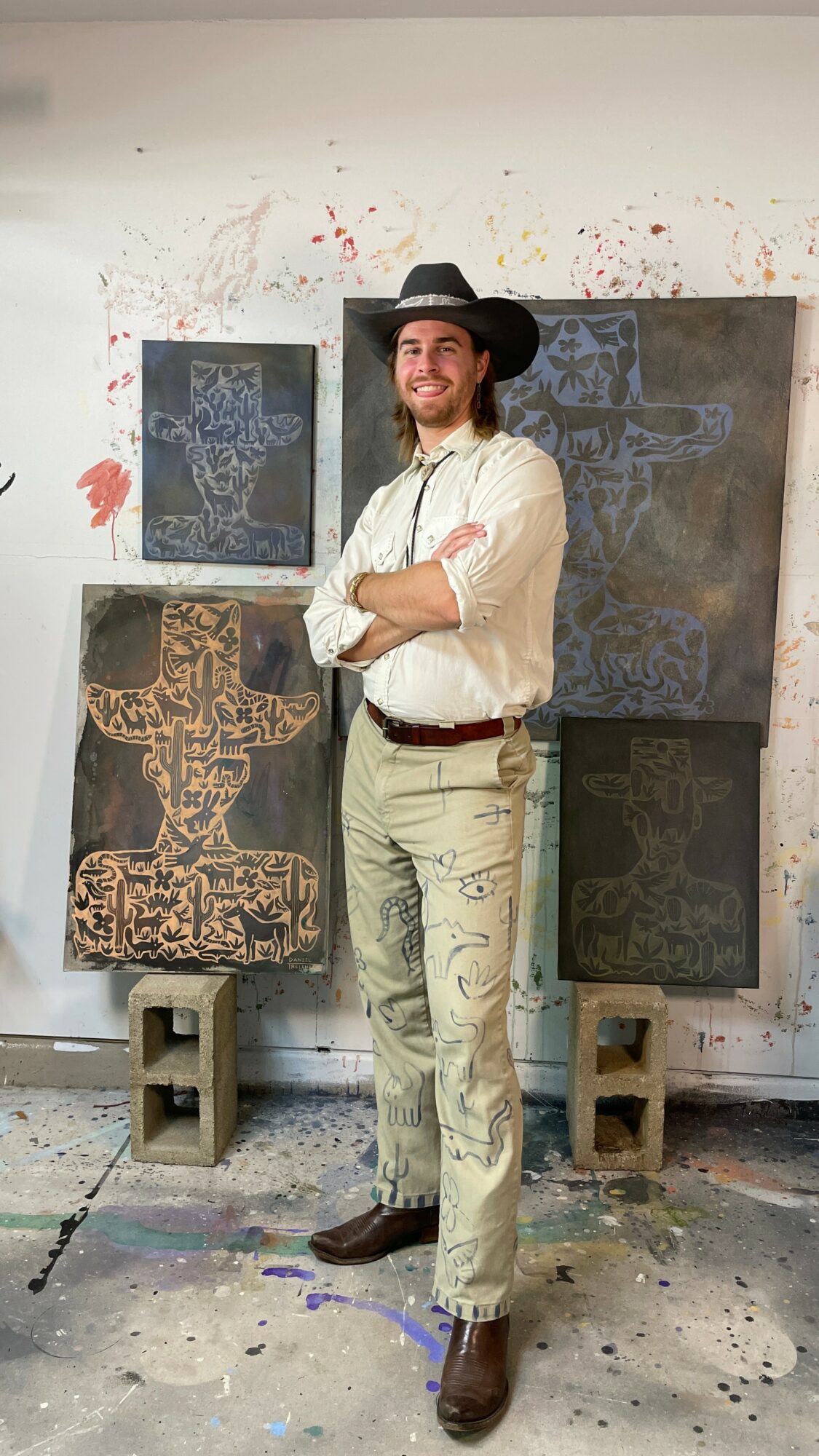 Man with a beard wearing a hat, white shirt, and beige pants standing in front of abstract artwork on a wall.