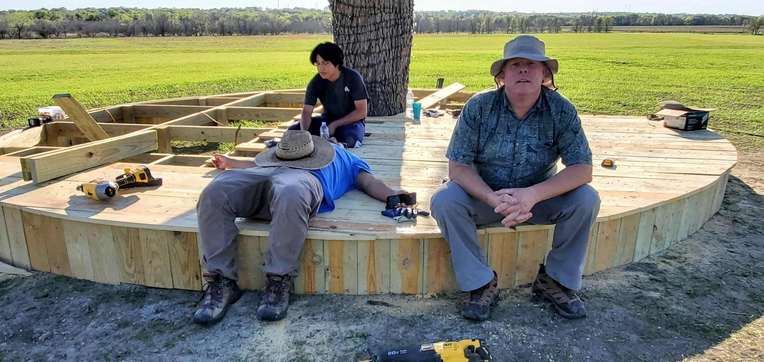 Three people on a wooden platform outdoors, with tools and a tree in the background, two sitting and one working.