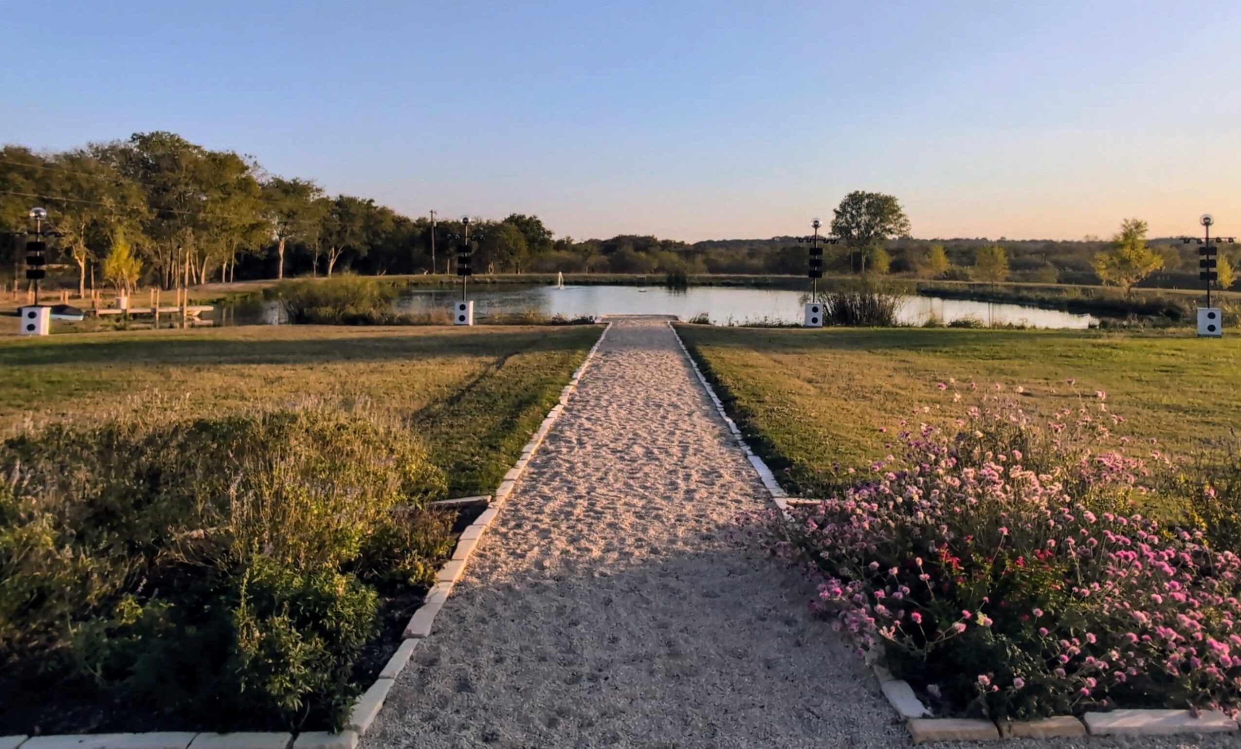 Path leading to a lake with trees and flowers on both sides, under a clear sky.
