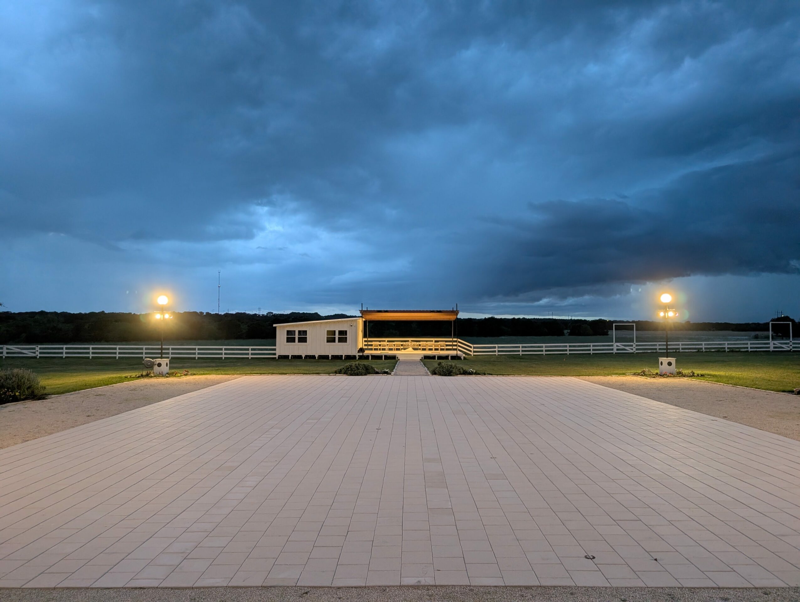 Wide view of an empty paved area with a building in the distance under a cloudy sky, two bright lights on either side.