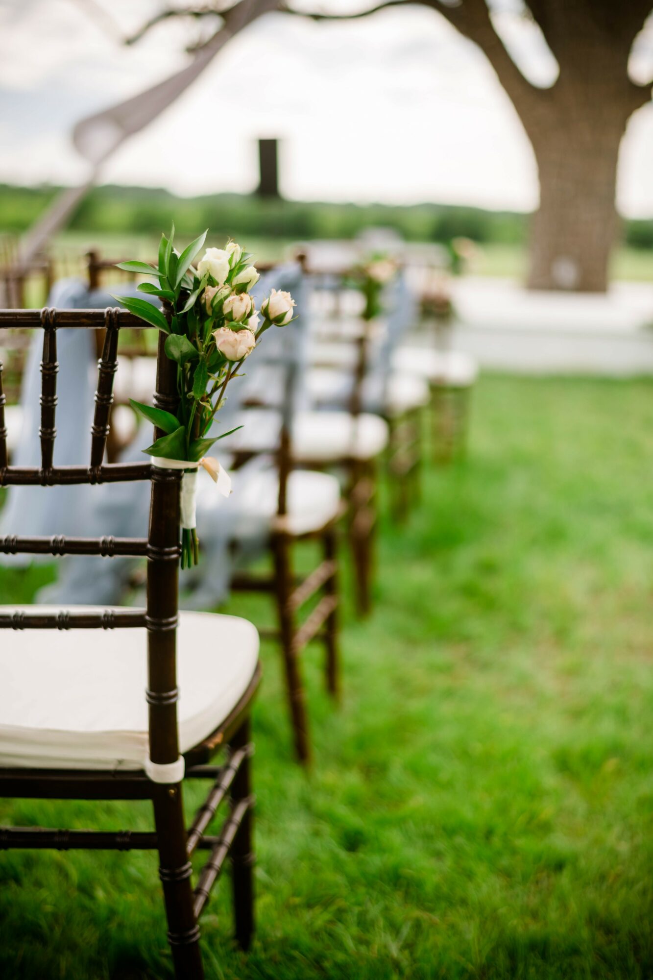 Chairs arranged outdoors with a floral decoration on the first chair, green grass, trees, and a body of water in the background.