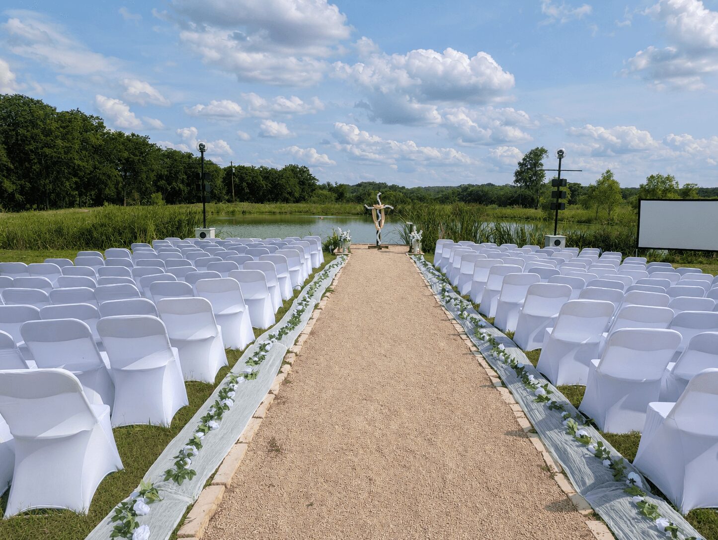 Outdoor wedding setup with chairs on both sides of a central aisle leading to an altar, under a partly cloudy sky.