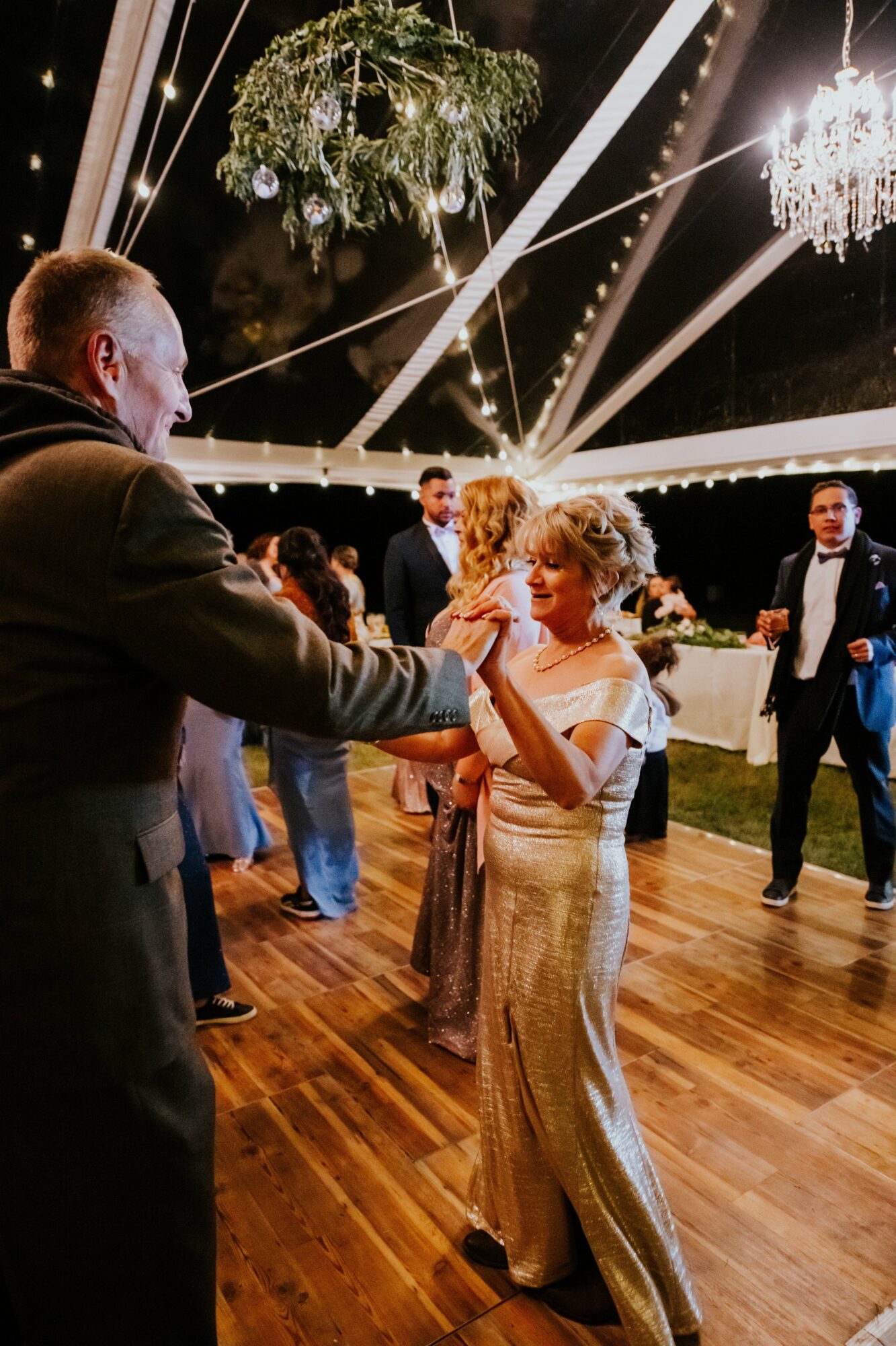 People dancing at an outdoor event with chandeliers and hanging plants, wooden floor, night setting, festive atmosphere.