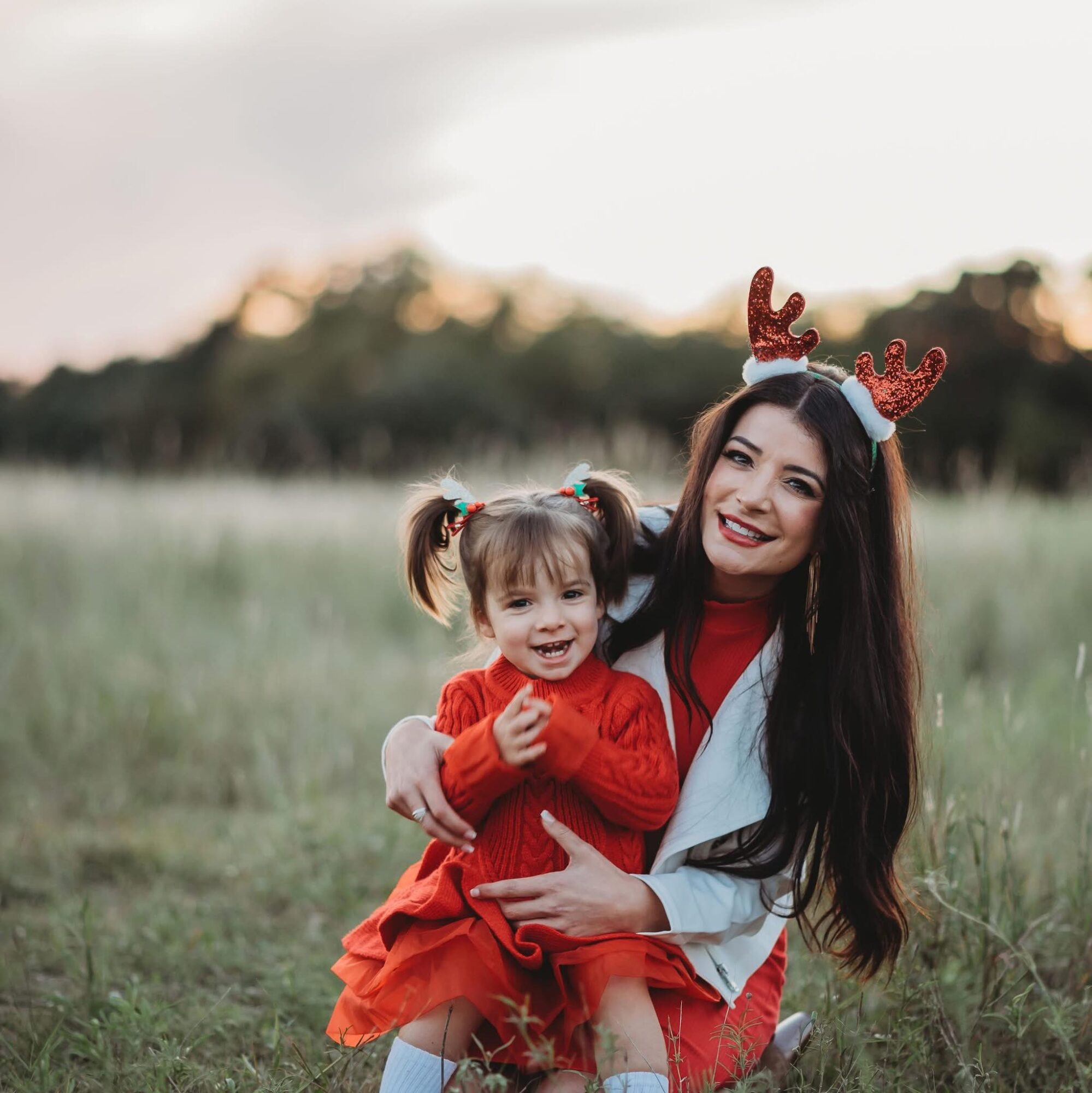 Woman and young girl outdoors, woman wearing reindeer antlers headband, girl in red dress, smiling, grassy field background