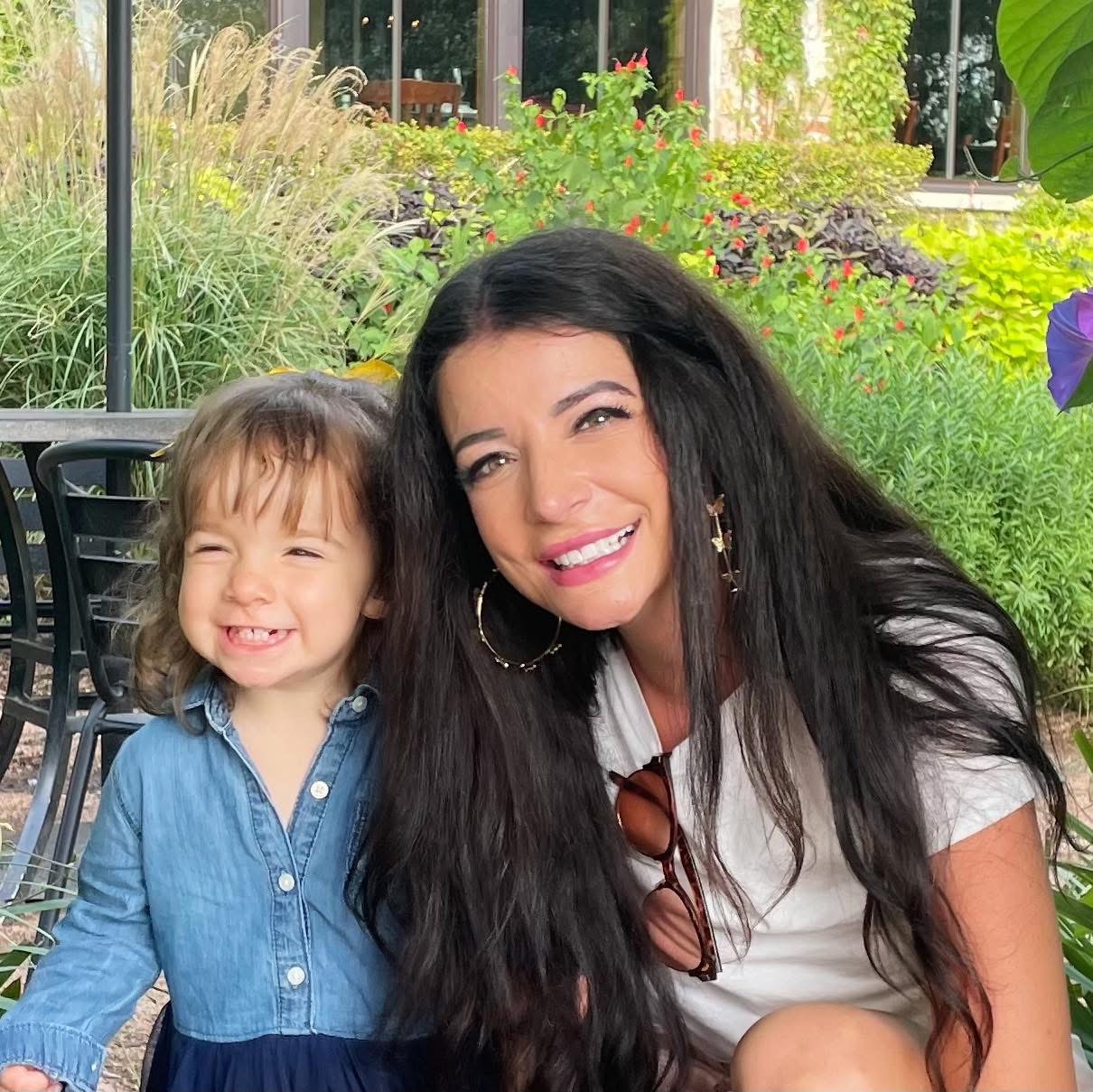 A woman and a young girl smiling outdoors with greenery in the background.