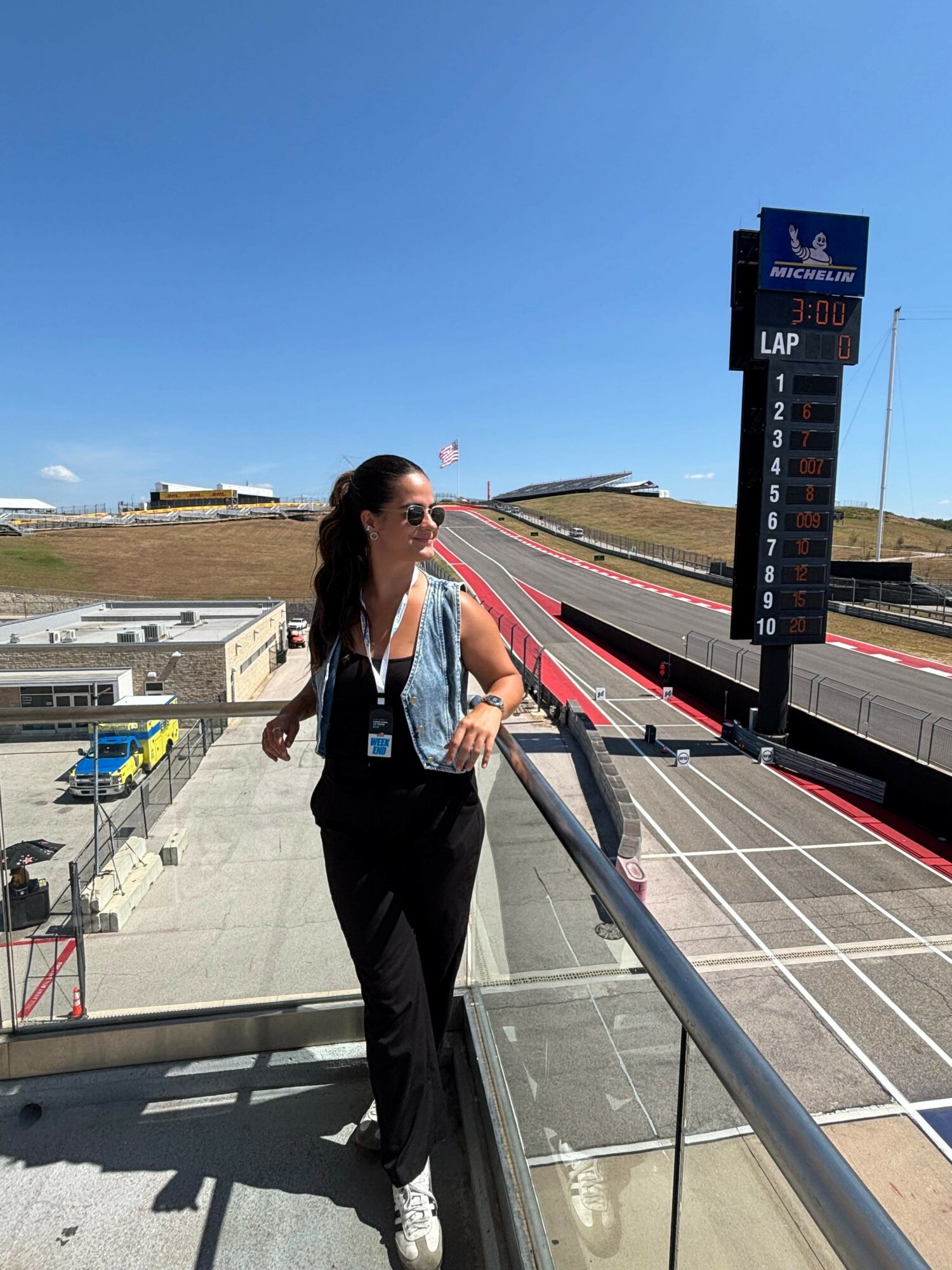 Woman with sunglasses and a backpack standing on a balcony overlooking a race track, clear blue sky, and a scoreboard.