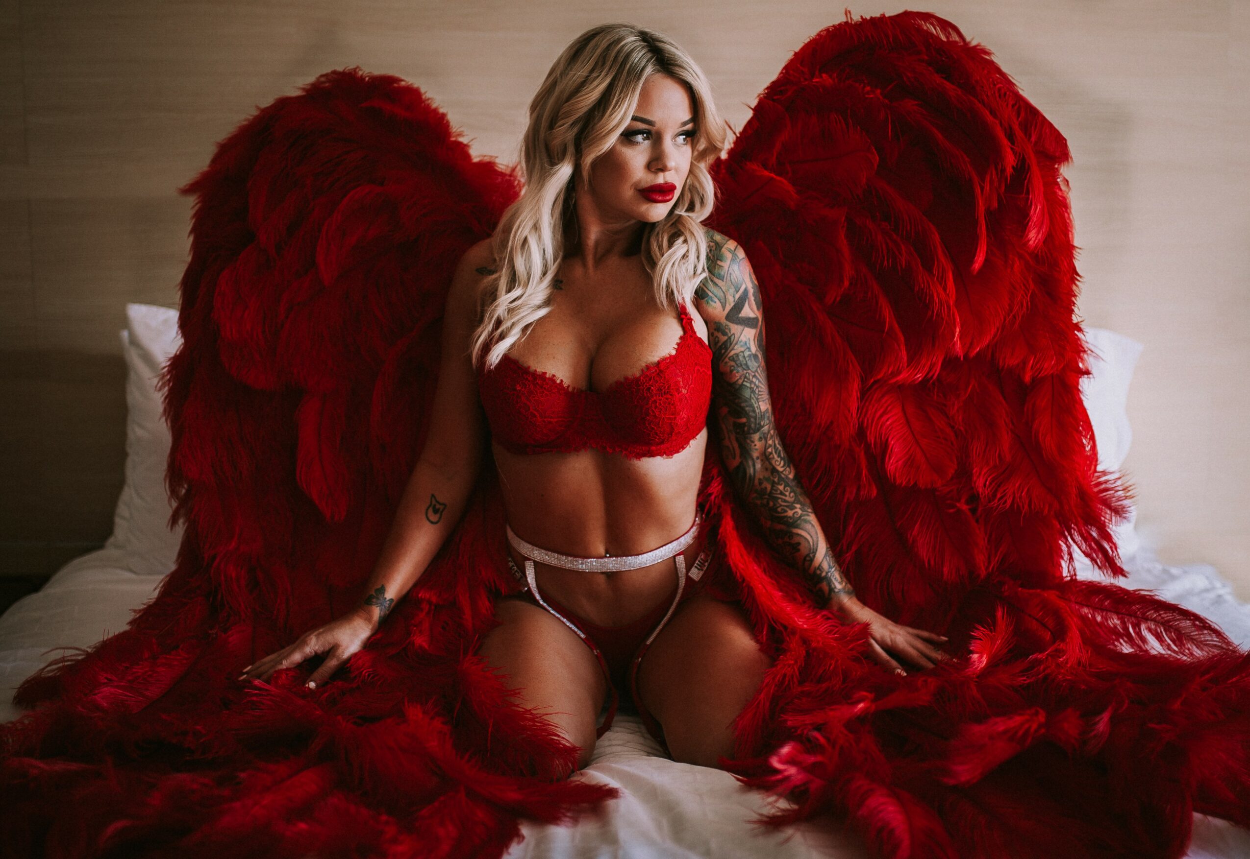 Woman with large red feathered wings, wearing red lingerie, sitting on bed with a neutral background.