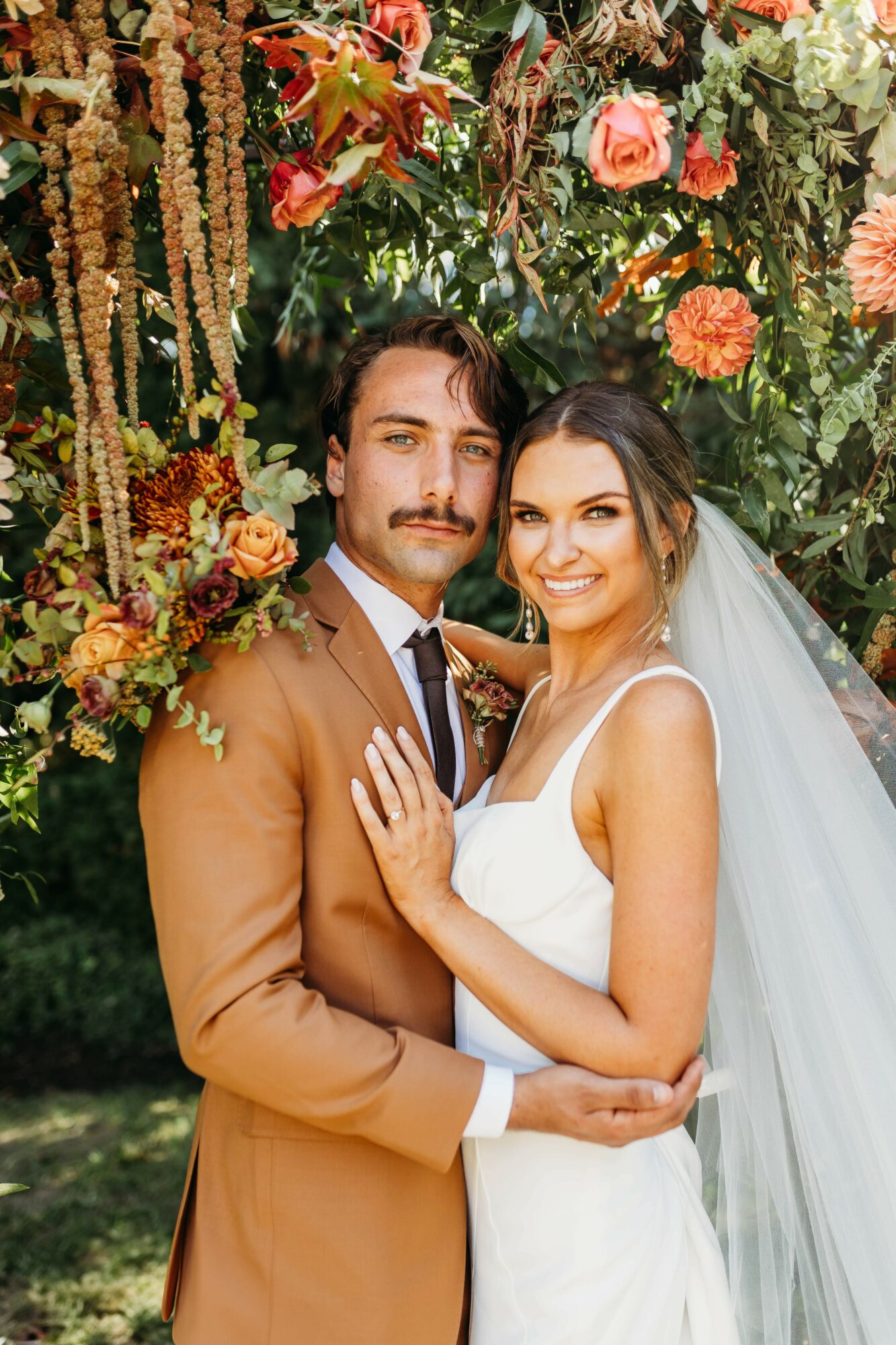 Bride and groom smiling, standing close, surrounded by colorful flowers and greenery, outdoors during daytime.