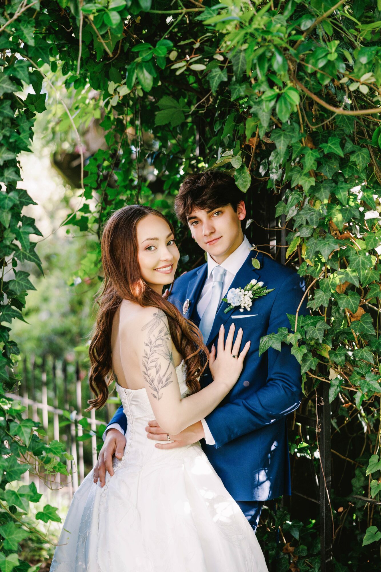 A young couple in formal attire standing close under a leafy tree, smiling at each other.