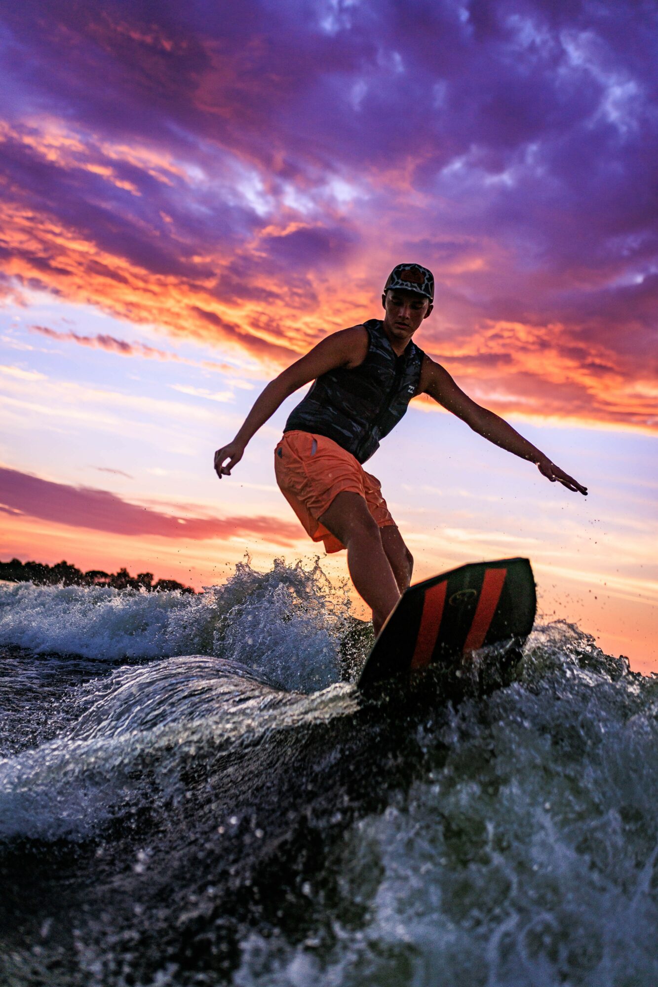 Person wakeboarding on water during sunset with colorful sky, wearing sunglasses, black vest, and orange shorts, balancing on board.