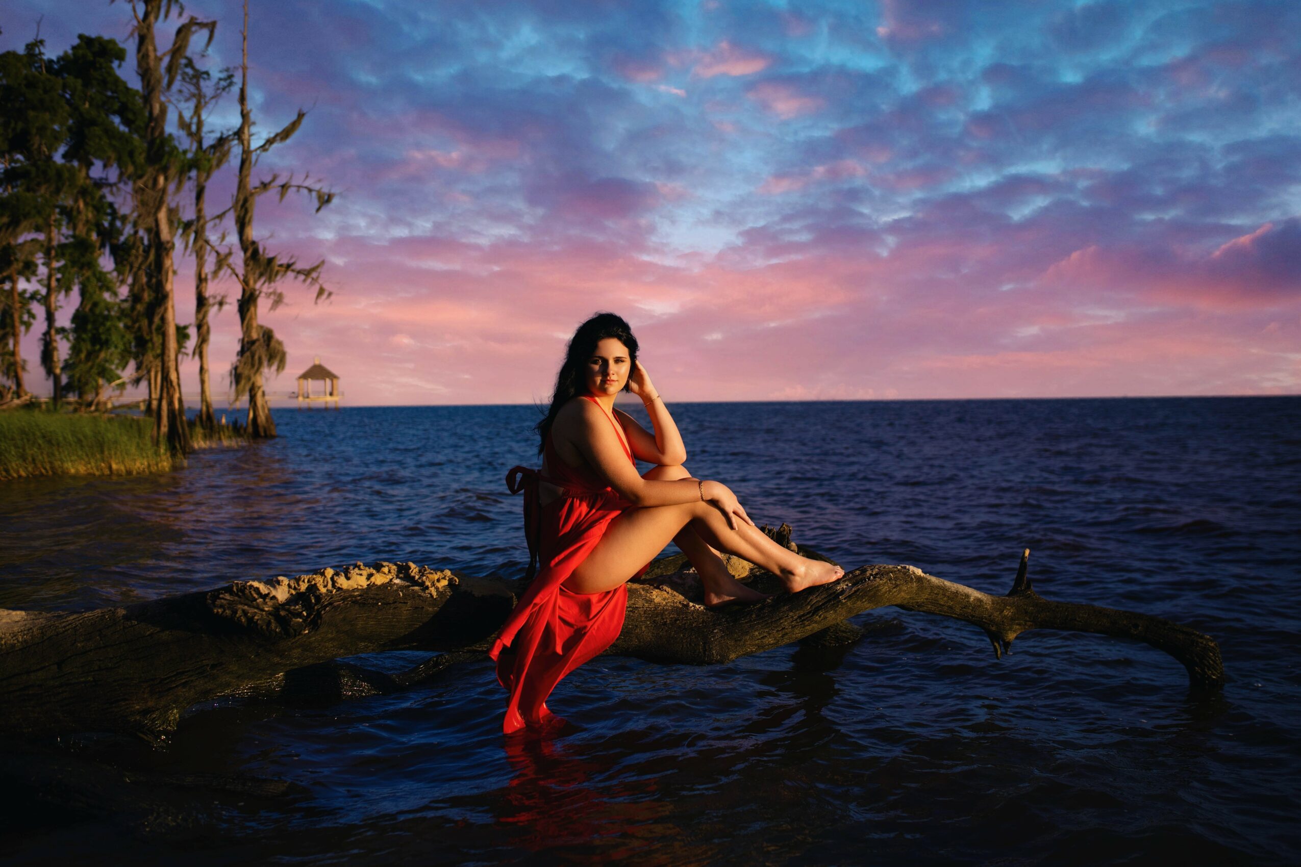 Woman in red dress sitting on a log by water during sunset, with trees and colorful sky in background.
