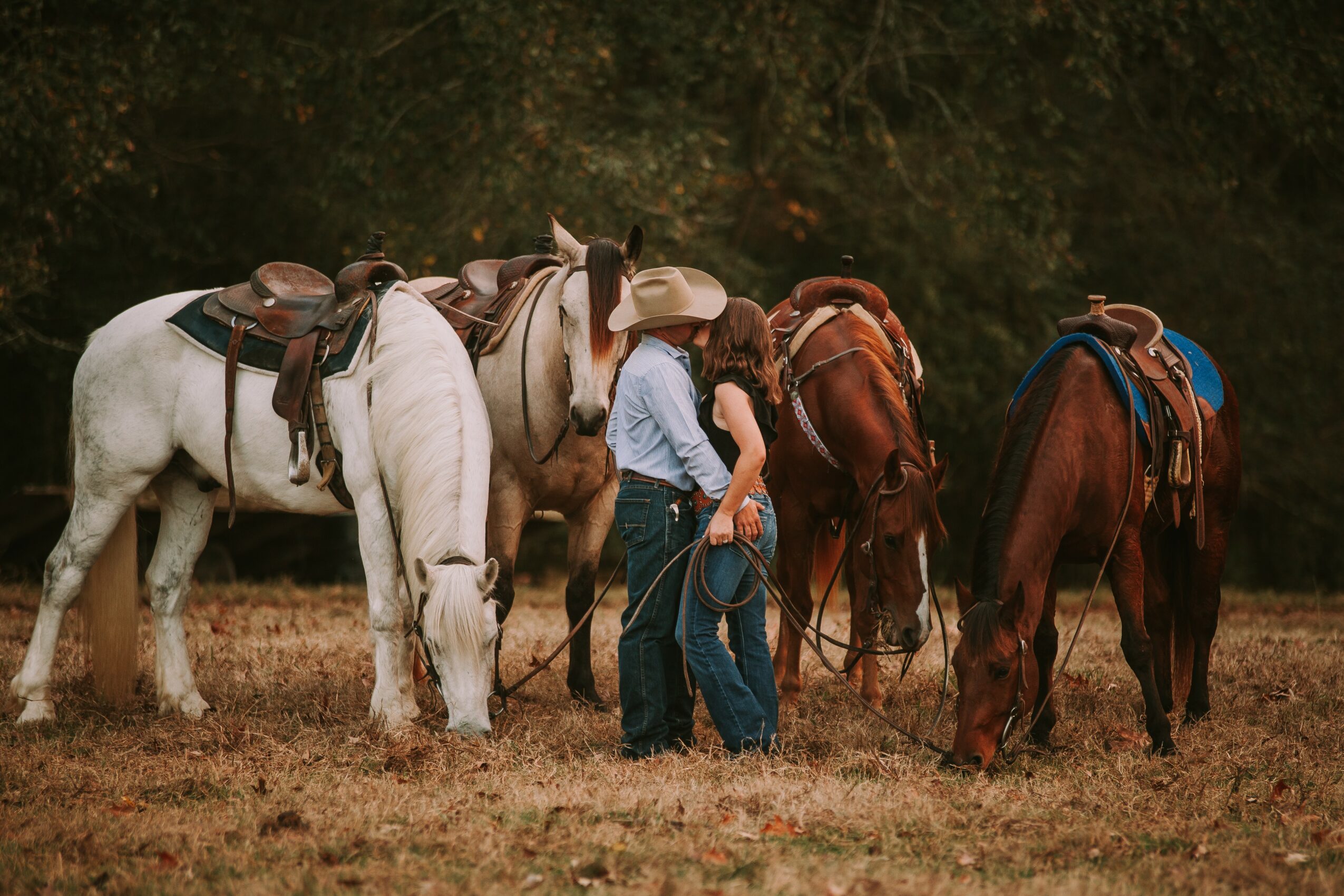 Person in cowboy hat and jeans stands among five horses in a field, with trees in the background.