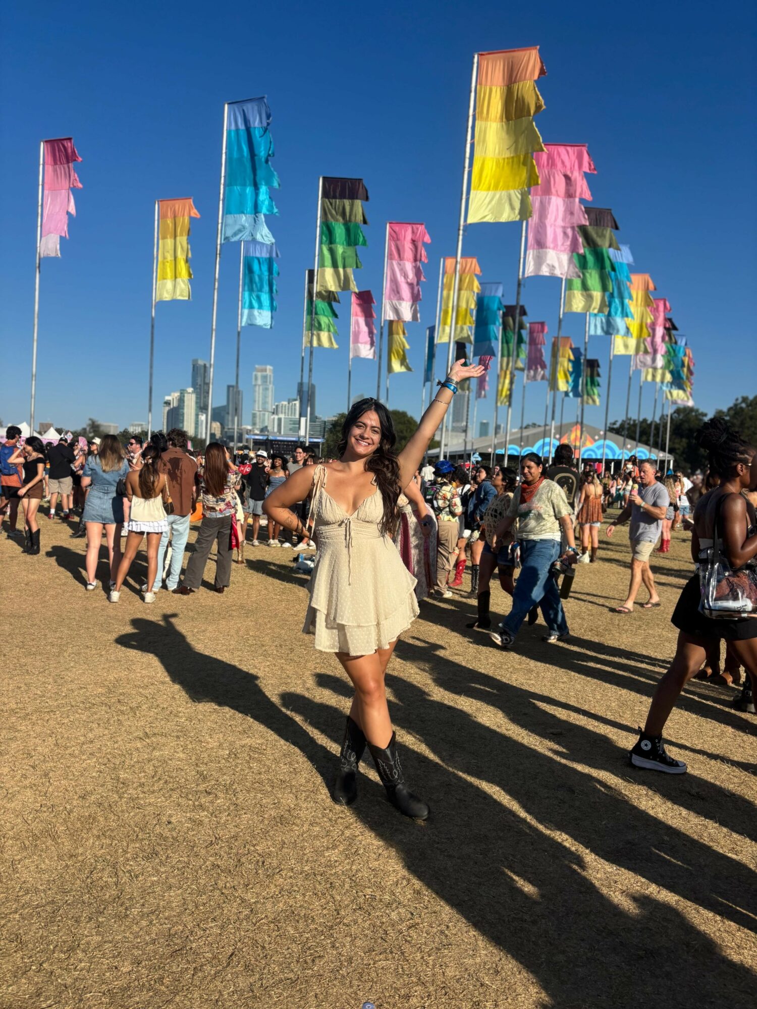 Woman in a yellow dress smiling and waving at an outdoor festival with colorful flags and a crowd, city skyline in background.