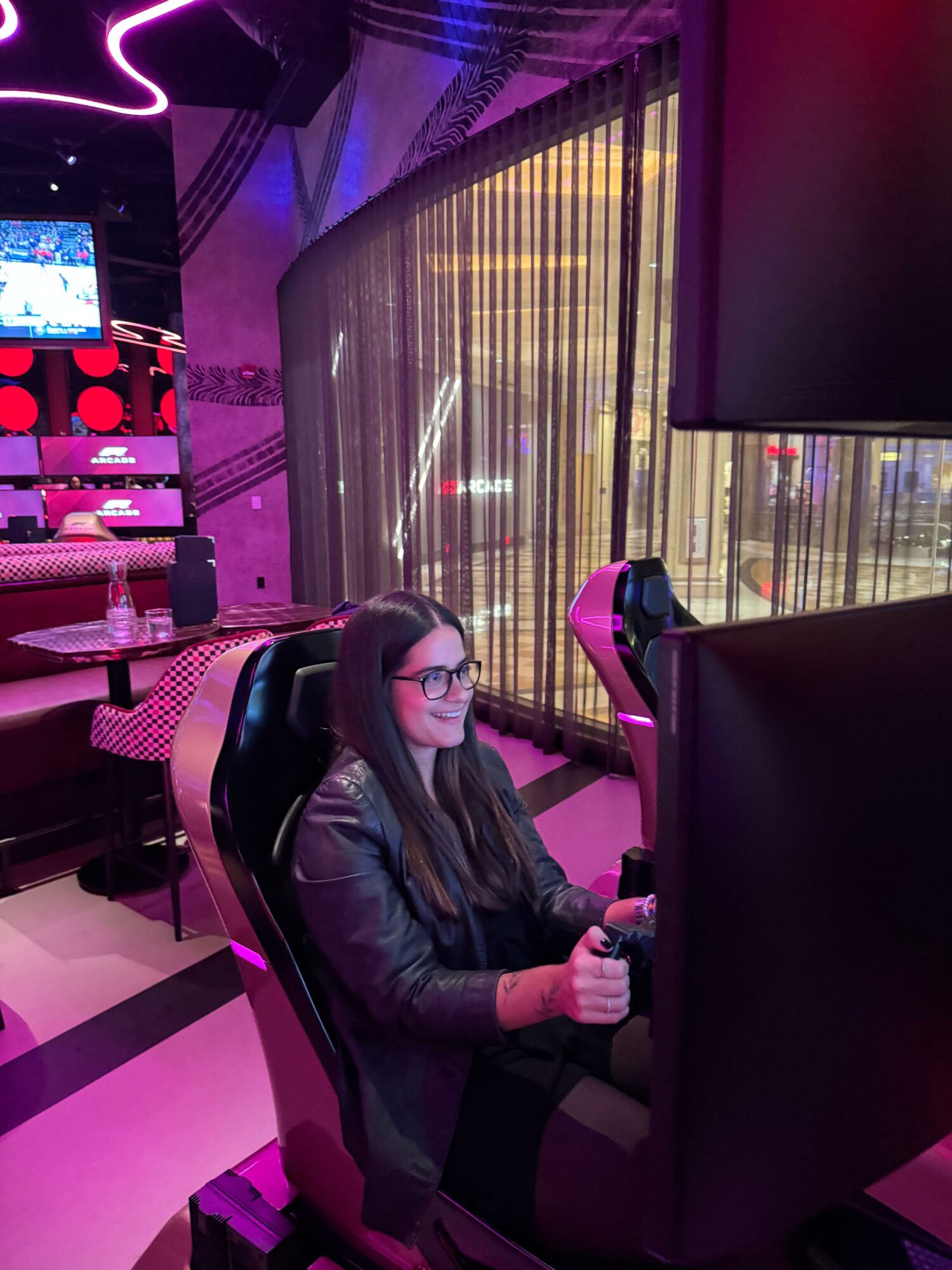 Young woman with glasses sitting in a gaming chair, focused on a screen in a colorful arcade or gaming area.