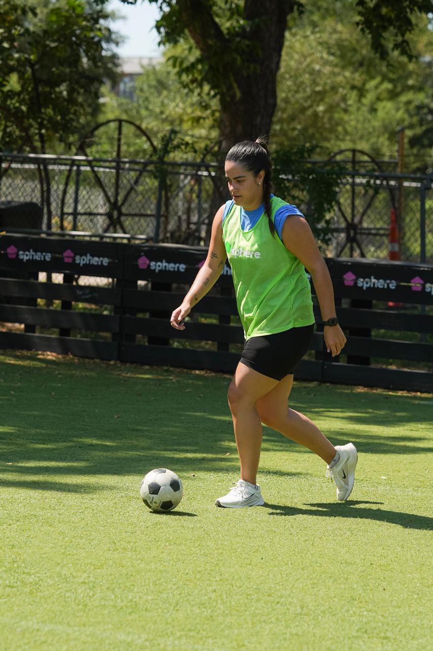 Woman in green vest and black shorts playing soccer on grass field, with trees and fence in background.