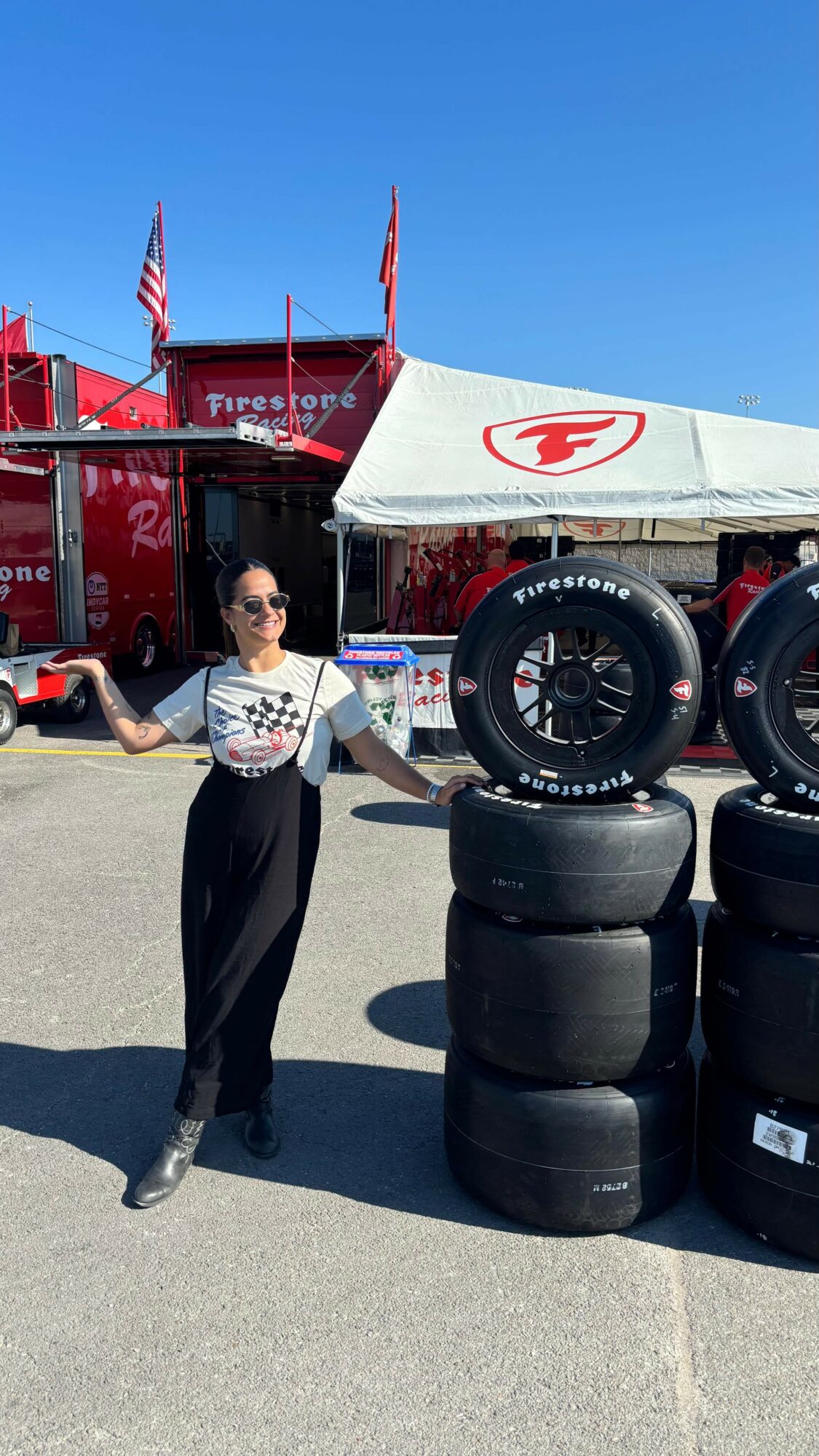Person standing next to stacked tires and a tent with a Firestone logo, outdoors on a sunny day.