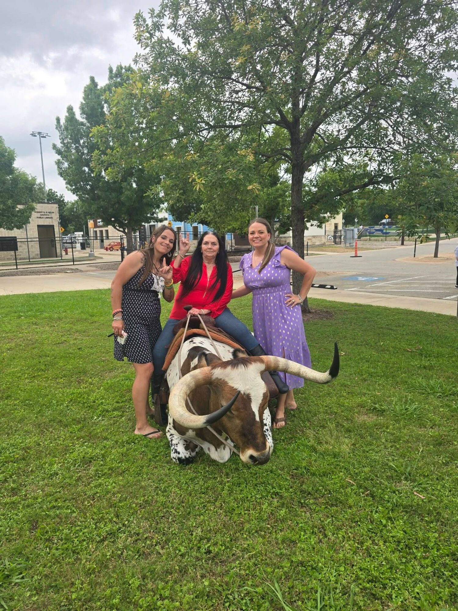 Three women standing on grass with a large cow lying down, trees and a parking lot in the background.
