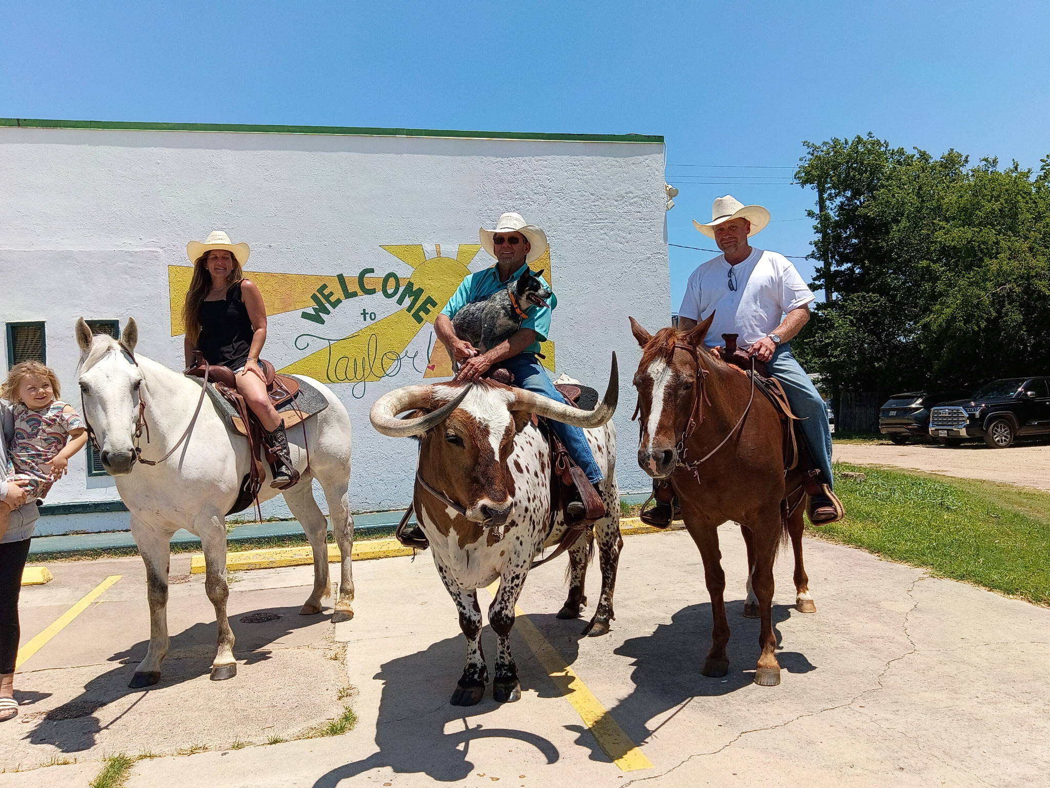 Four people on horseback in front of a white building with a welcome sign, sunny day, trees in background.