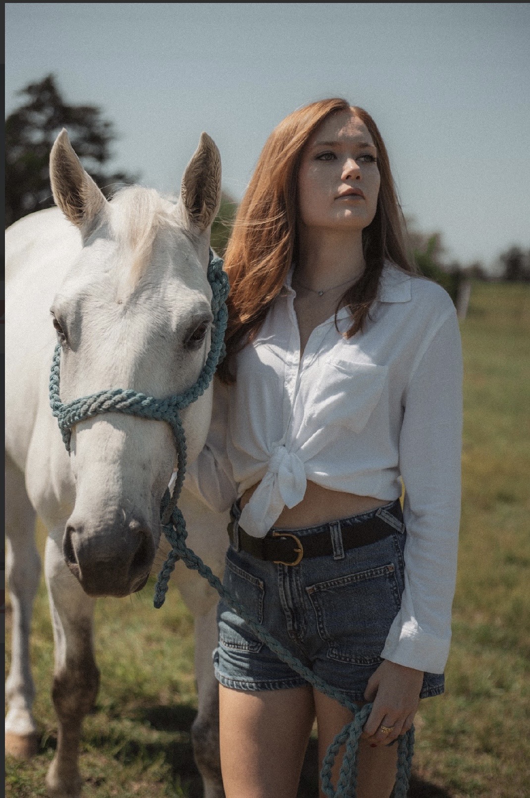 Young woman with long red hair standing next to a white horse outdoors, wearing a white shirt and denim shorts.