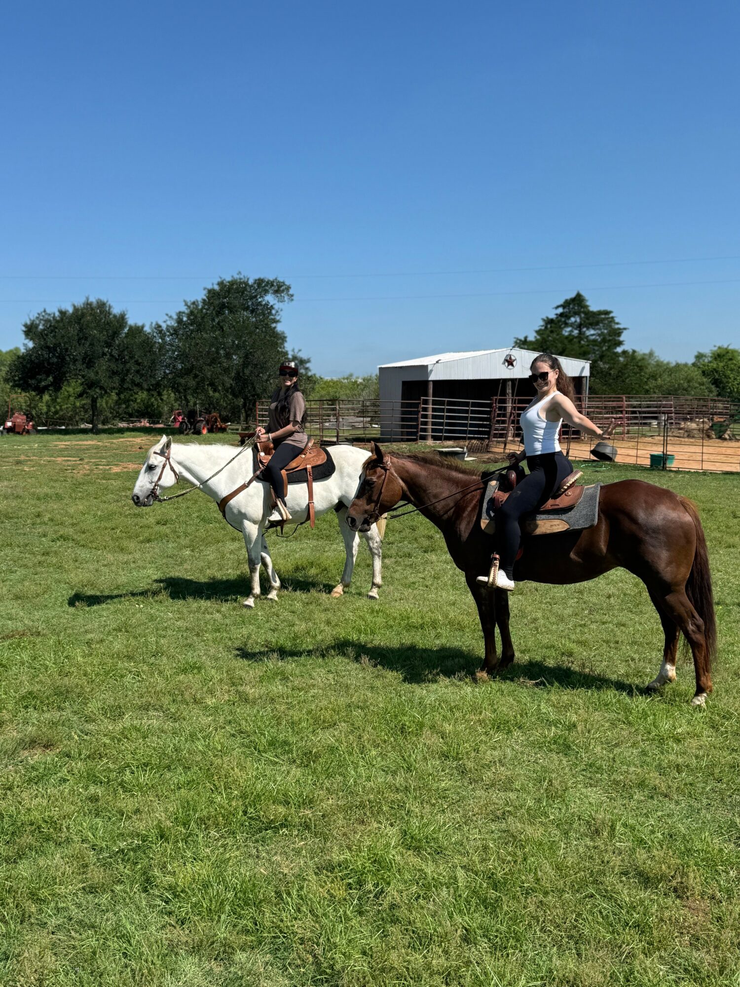 Two women riding horses in a grassy field with trees and a building in the background.