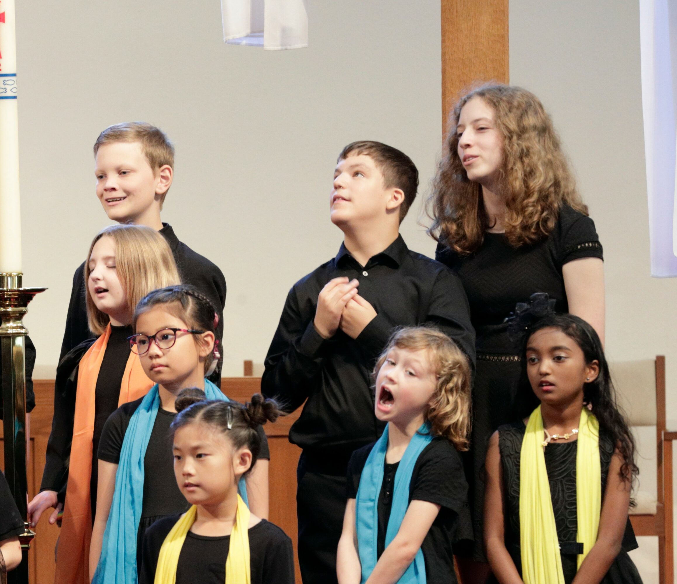 Group of children and a young woman standing on stage, some singing or speaking, with a plain background.