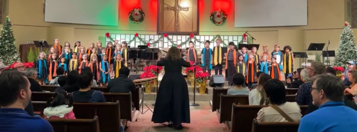 Choir performing on stage in a church with audience seated, Christmas decorations, and a conductor in front.