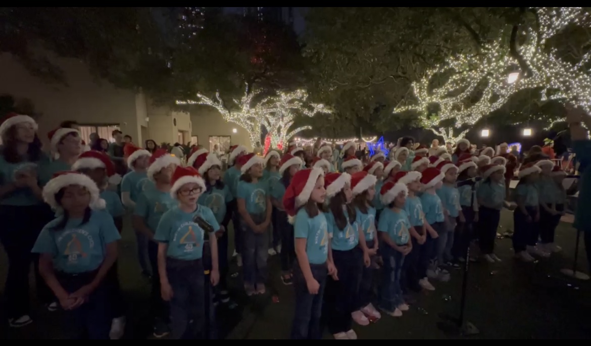 Children wearing Santa hats and blue shirts gather outdoors at night with festive lights overhead.