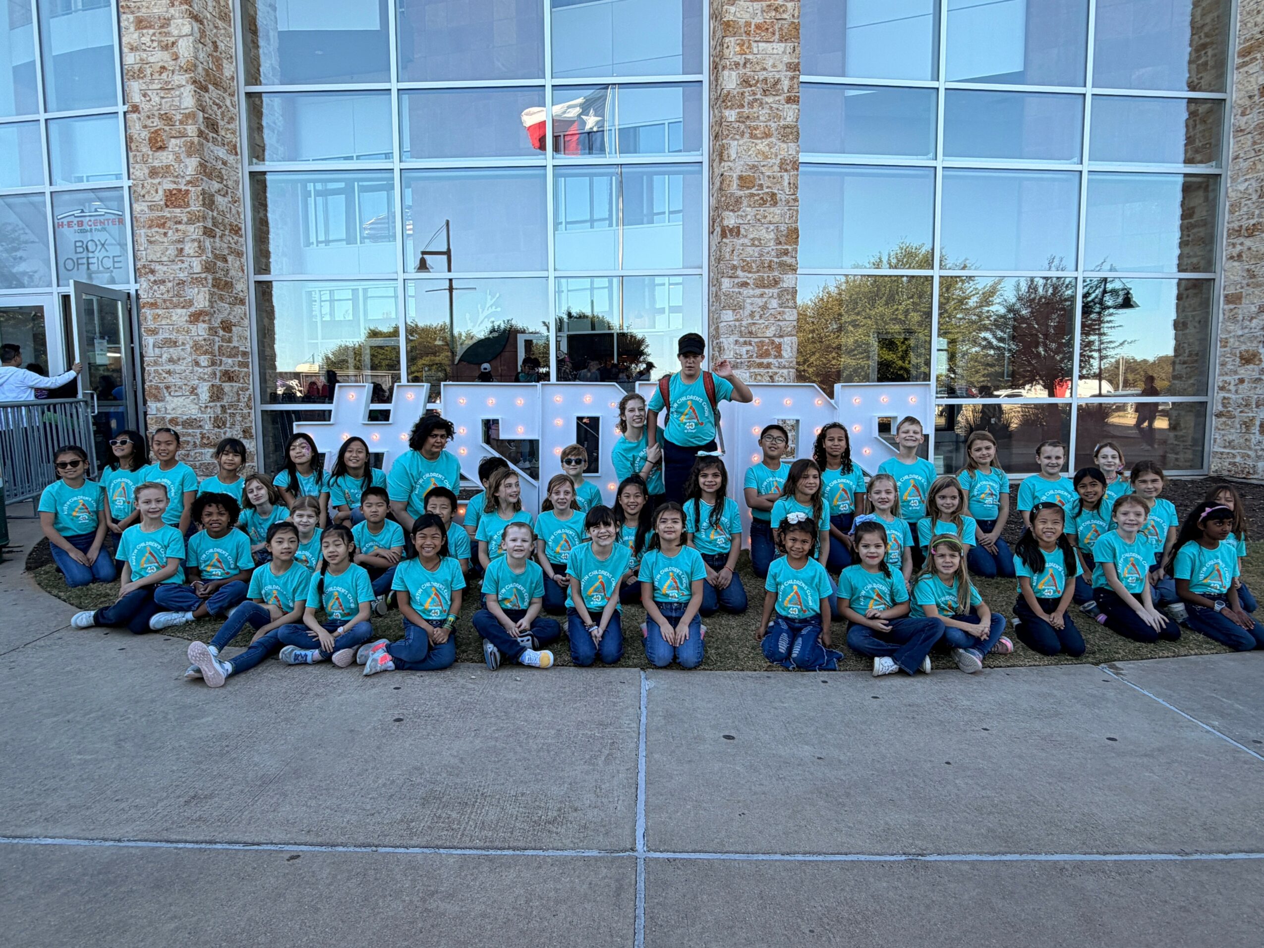 Group of children and adults in matching blue shirts outside a building with large glass windows, posing for a photo.
