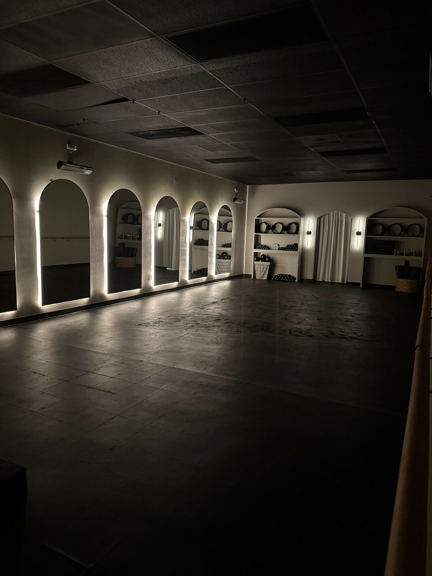 Long corridor with arched doorways and illuminated interior spaces, dark flooring, and ceiling, viewed from the side.