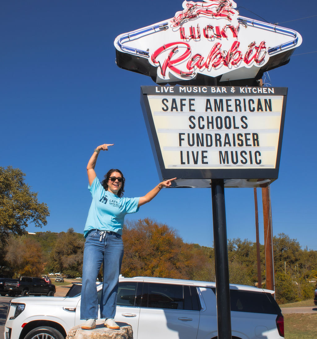 Person standing next to a sign for Lucky Rabbit, pointing at the sign, with trees and cars in the background.