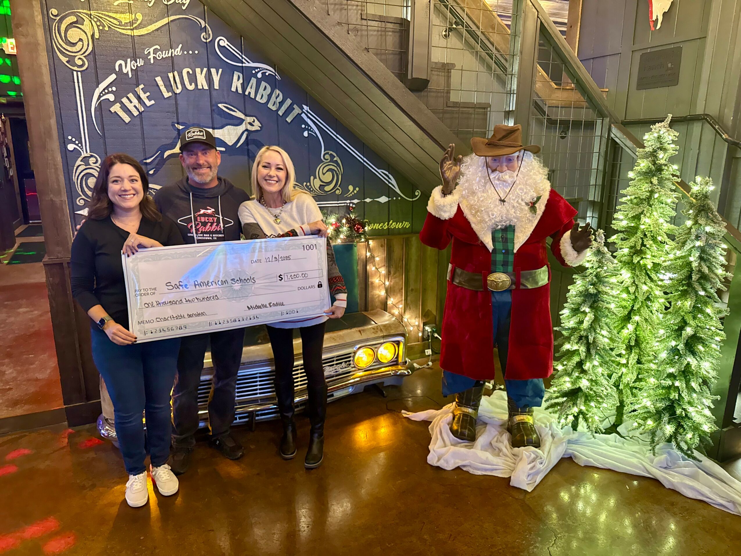 Four people stand indoors holding a large check, with a Santa costume and Christmas trees nearby, in a festive setting.