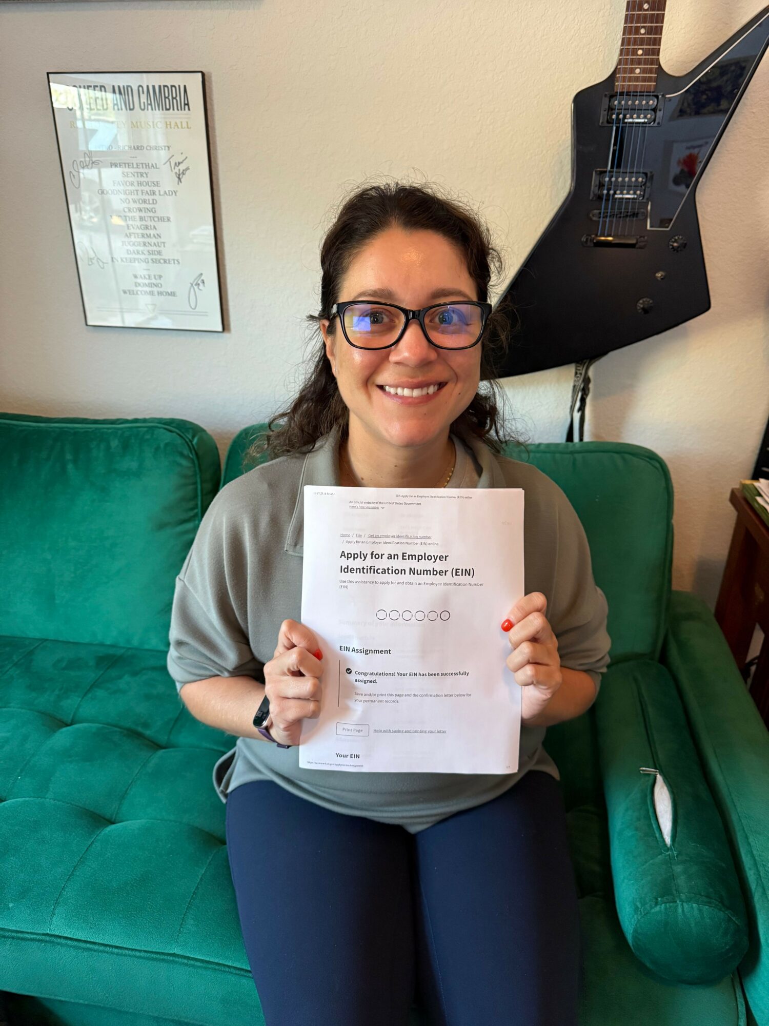 Woman with glasses holding a document, sitting on a green couch, smiling, in a room with a guitar and framed certificate.