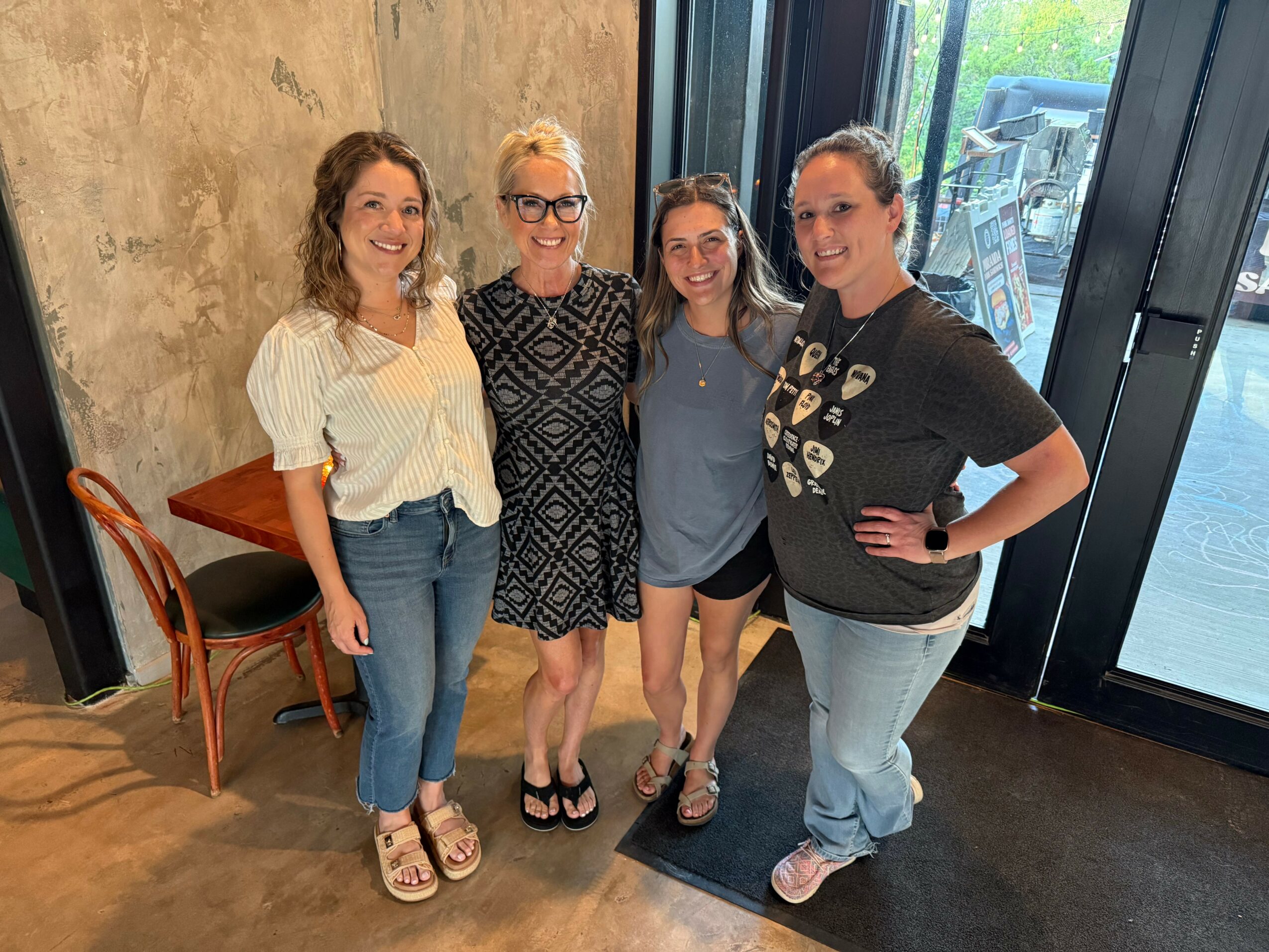 Four women standing together indoors near a large window, smiling, with a concrete wall behind them.