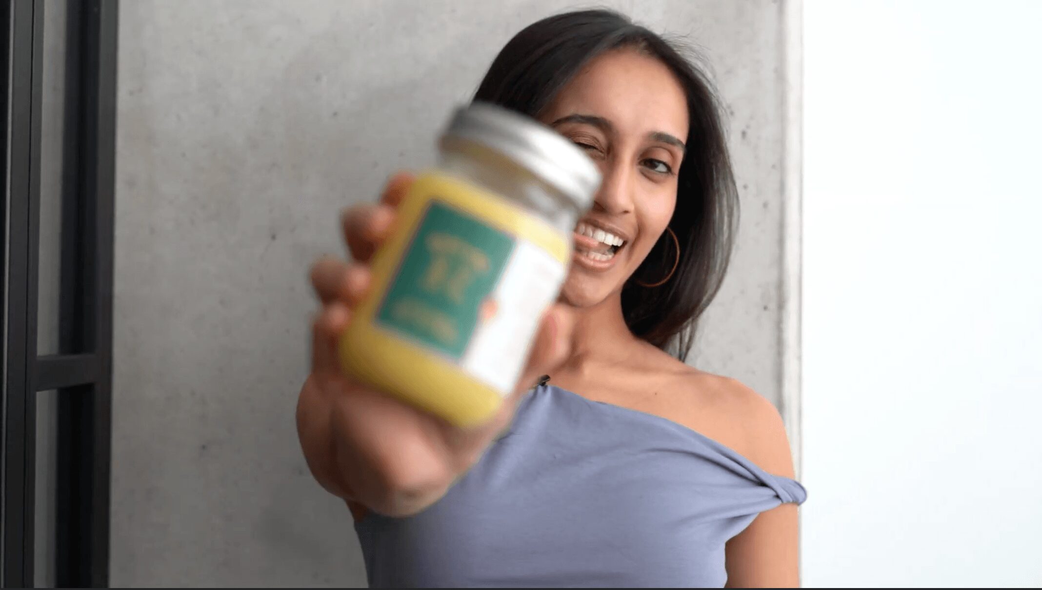 Smiling woman holding a jar towards the camera, with a concrete wall background.
